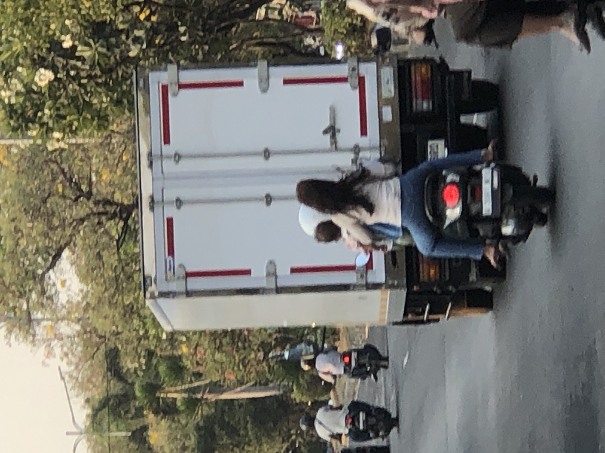 In Ngaban, Indonesia, a truck carrying a white refrigerated box is seen on a road with trees lining the sides. Multiple motorbikes are present. In the foreground, a woman with long dark hair is riding as a passenger on a motorbike, wearing a light-colored shirt and blue jeans. A child is also on the motorbike with her. To the left, two men are riding motorbikes, both wearing helmets. The overall scene depicts traffic on a street in a city, likely during daylight hours, with a focus on the movement of vehicles and people.