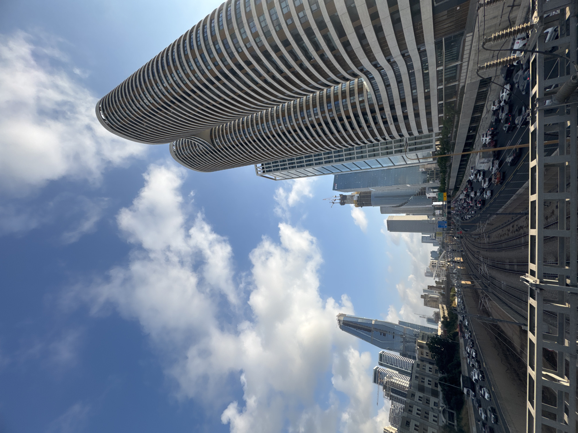 The image displays a dynamic urban landscape with a focus on a unique architectural structure that appears to be two large, curved buildings. These buildings are characterized by horizontal white and beige bands that create a wavy, organic form. In the background, a dense collection of modern skyscrapers rises against a bright blue sky dotted with white, fluffy clouds, suggesting a clear and sunny day.
To the right of the buildings, a multi-lane road is visible with a significant amount of traffic. Cars are densely packed, indicating a traffic jam. Adjacent to the road are railway tracks, also curving through the urban environment. Power lines and supporting structures are prominent in the foreground on the right side of the image, framing the scene.
The perspective is from below, looking up at the curved buildings and out towards the city skyline and infrastructure. The lighting suggests it is daytime, likely mid-morning or afternoon, given the clear sky and bright conditions. There are no people clearly visible in the image, but the presence of cars implies human activity. The scene captures the bustling energy of a modern city, with a mix of striking architecture, transportation networks, and commercial development. Some faint text is visible on one of the buildings in the background, appearing to be part of the facade. The overall impression is one of a vibrant and active urban center under a pleasant sky.