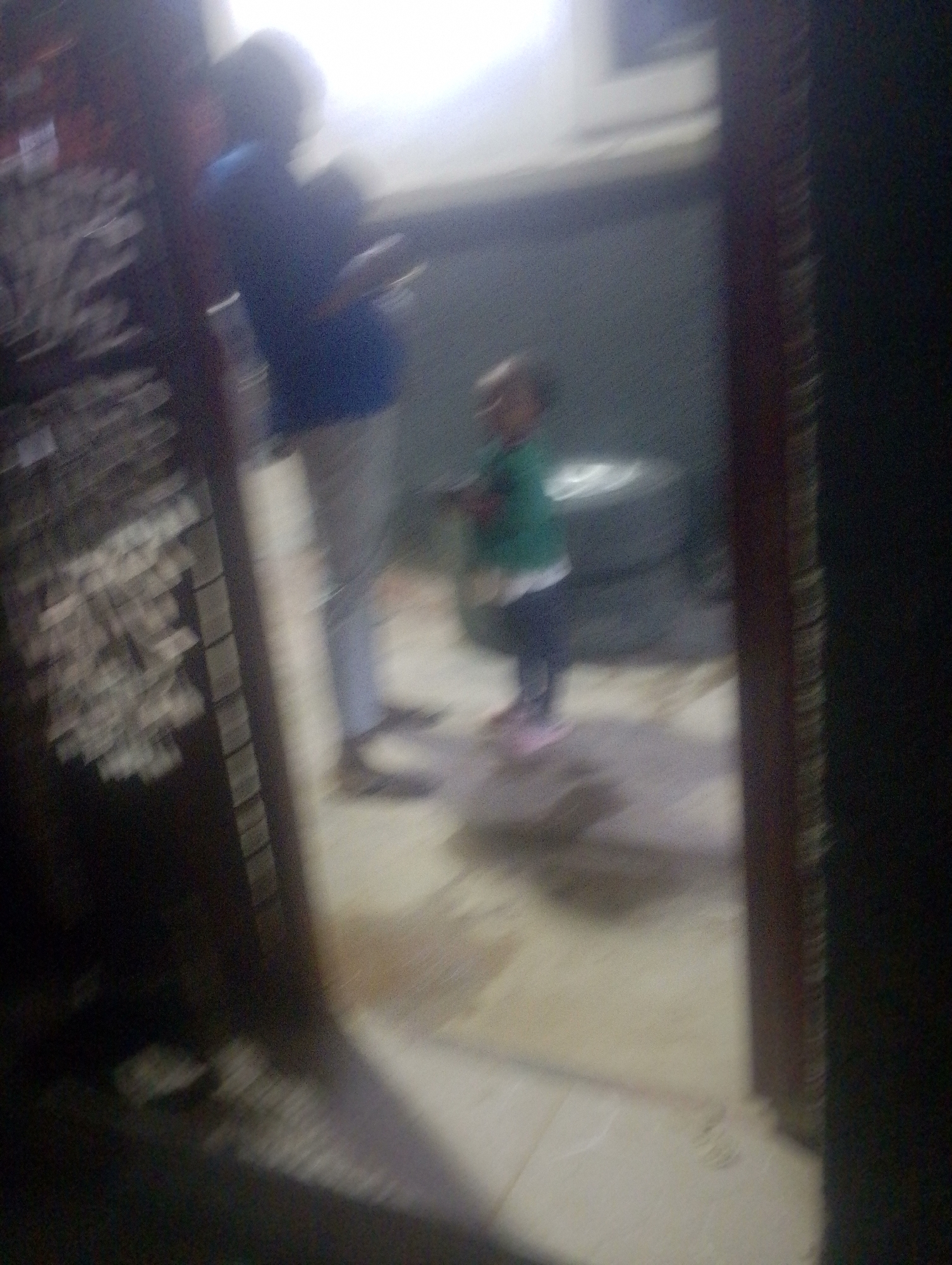 In the city of Jos, Nigeria, an out-of-focus image captures two individuals in an interior space. The primary subject, a child, stands in the center, wearing a green shirt and dark pants, with pink shoes. To the left, a taller, blurred figure in a dark blue shirt and light pants is also visible. The background appears to be a wall with a dark, circular object, possibly a tire, behind the child. The floor is tiled in a light color. The overall lighting is bright and diffused, suggesting an indoor environment with a light source from the upper left. The image is characterized by motion blur, making precise details difficult to discern.