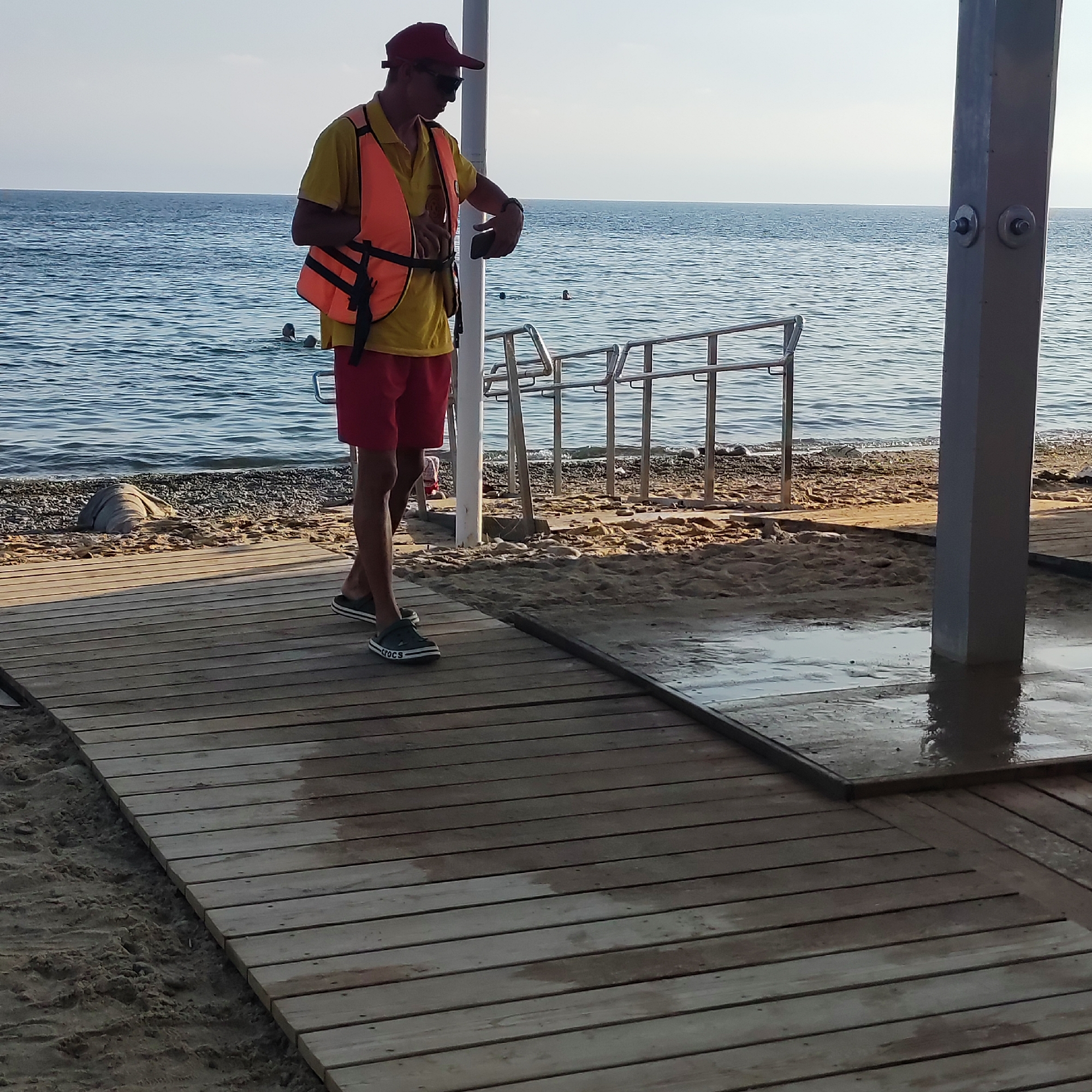 A lifeguard wearing a red cap, sunglasses, yellow shirt, and orange life vest stands on a wooden boardwalk by the sea. He is looking down at his phone. Red shorts and dark sandals complete his attire. The sea is calm with a few swimmers in the distance. To the right of the boardwalk is a beach with sand and pebbles, and a silver shower fixture.