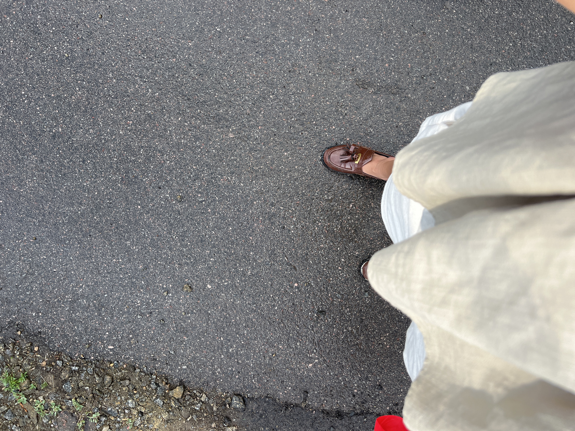 A first-person, top-down perspective image shows a person standing on a dark gray asphalt surface. The person is wearing a long, light-colored dress and a brown leather loafer with tassels on the right foot. The asphalt shows signs of wear with small stones embedded in the surface. The bottom left of the frame includes dirt and small rocks along the edge of the asphalt. No identifying information indicates the location as Boguchar, Russia.