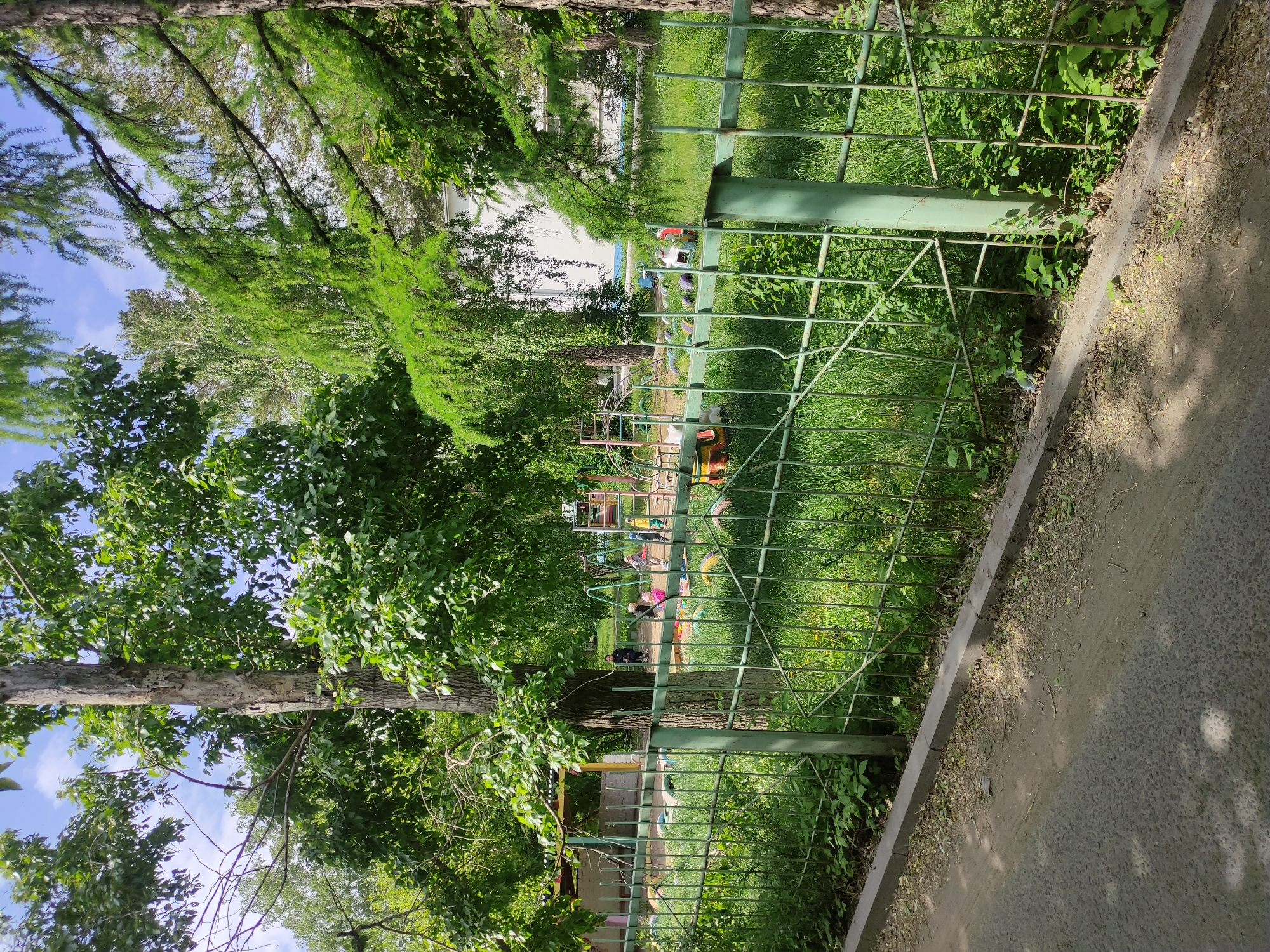 The image shows a playground located in Pavlodar, Kazakhstan. A green metal fence separates the playground from a concrete sidewalk. Lush green grass and trees surround the playground. In the background, playground equipment and several people, including children, are visible. The weather appears to be sunny and clear.
