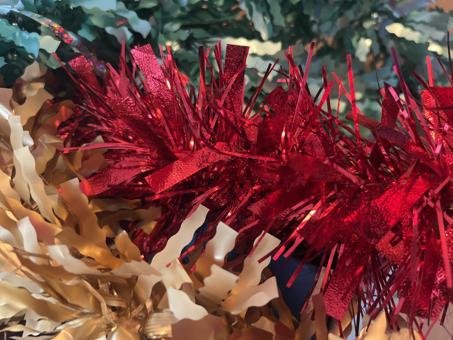 This close-up image depicts a dense arrangement of festive decorations, located in the city of Callbach, Germany. The central and lower-right areas are dominated by bright red tinsel, composed of narrow, reflective, metallic strips. Interspersed within the red tinsel are small, indistinct points of light. To the upper left, a section of gold-colored garland is visible, featuring wider, wavy metallic strips with a matte to semi-gloss finish. On the far right, a blurred green element, consistent with artificial evergreen foliage or green tinsel, provides a background contrast. No individuals, actions, or specific structural elements of a room are discernible; the focus is solely on the interwoven decorative materials.