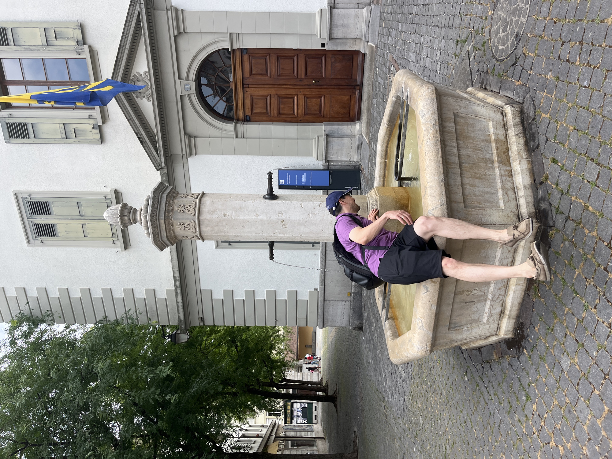 In the city of Vevey, Switzerland, on a cobblestone square, a person is crouched by an octagonal stone fountain, reaching into the water. The individual wears a purple t-shirt, black shorts, a blue baseball cap, and headphones around their neck. A black backpack is slung over their shoulders. To the left of the fountain, a large tree with green foliage stands beside a white building with grey decorative elements. The building features shuttered windows and a prominent arched wooden doorway. A blue and yellow flag hangs from a window above the doorway. In the background, a street with shops and more trees is visible, with several people walking in the distance.