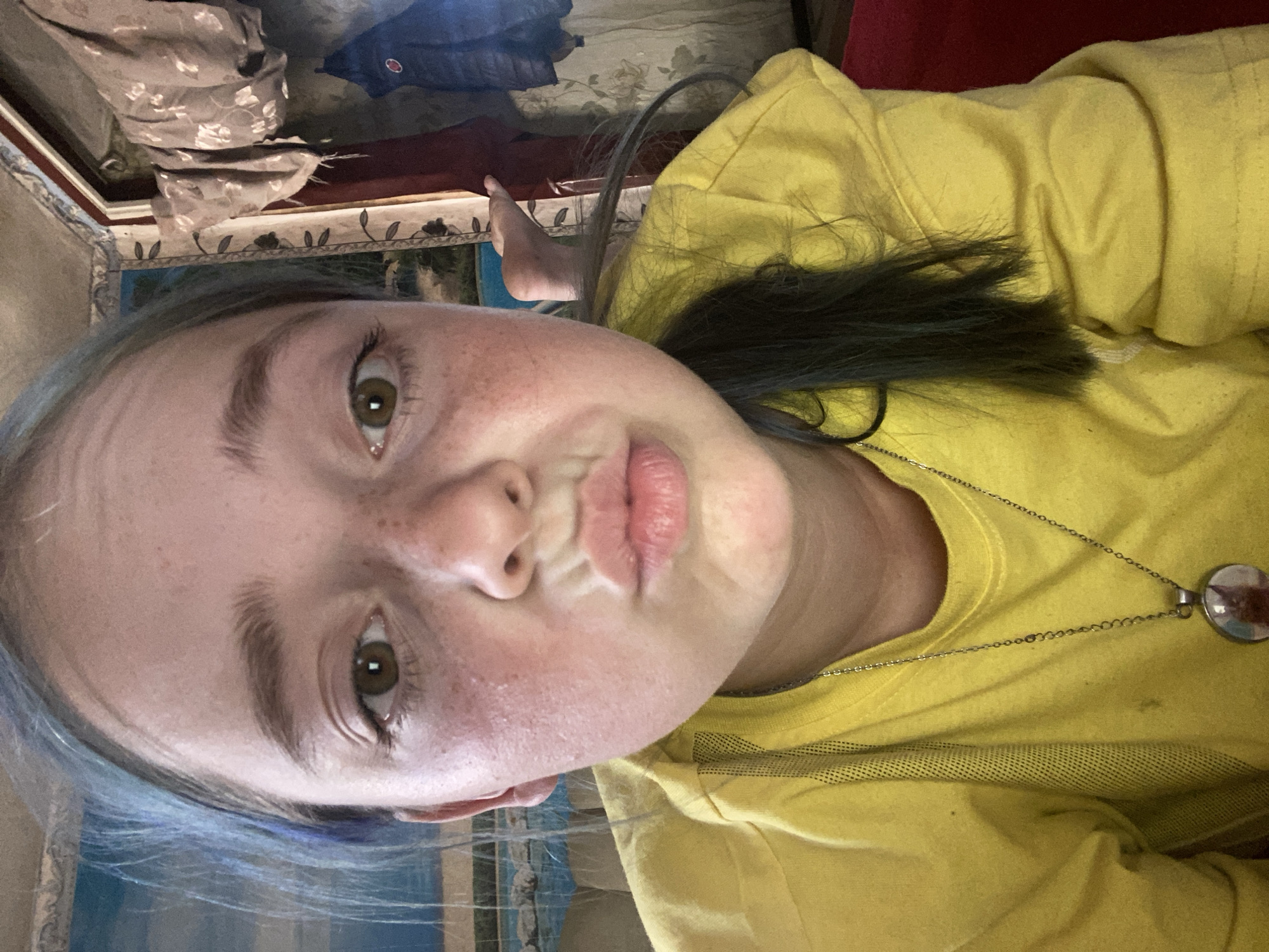 The image shows a close-up, front-facing selfie of a young person with light skin, freckles, and blue-tinted hair. The person is wearing a bright yellow t-shirt with a necklace featuring a circular pendant. Their lips are pursed, and their eyes are open and looking at the camera. The background is blurred but appears to be an indoor setting with curtains and what looks like a landscape painting on the left. There is no identifiable information within the image that suggests the location is in Nova Praha, Ukraine.
