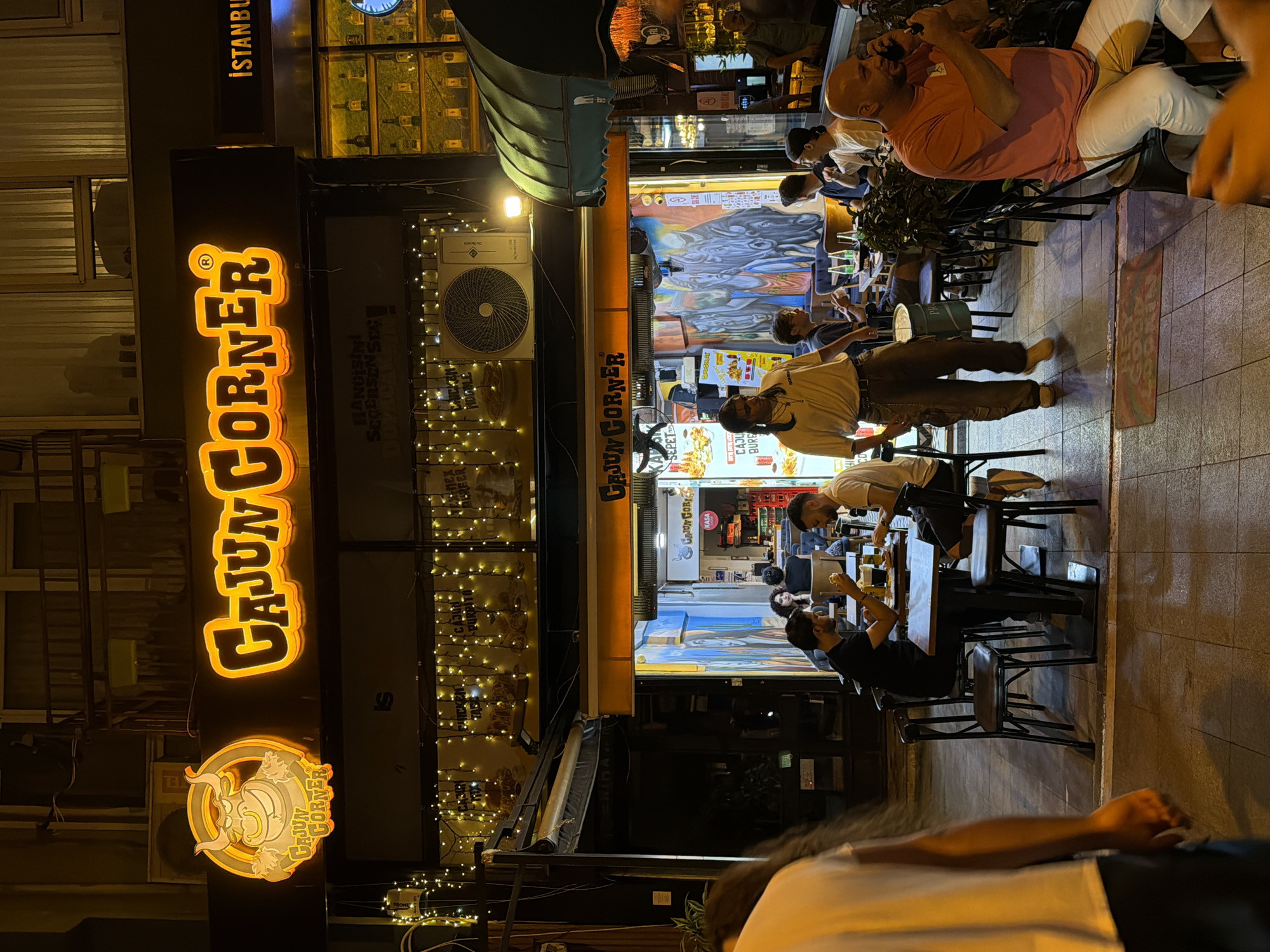 The image depicts the exterior of a restaurant named "Cajun Corner" located in Kadıköy, Turkey. The restaurant has a prominent neon sign featuring the restaurant's name and a cartoon bull mascot. Multiple people are seated at tables outside the restaurant, some of whom appear to be dining. The environment is lit by artificial lights, creating a warm and inviting ambiance. A person in a light-colored shirt is walking past the tables, and another person in a pink shirt is seated on a stool and appears to be on a phone call. The overall scene suggests a lively evening at a popular dining spot.