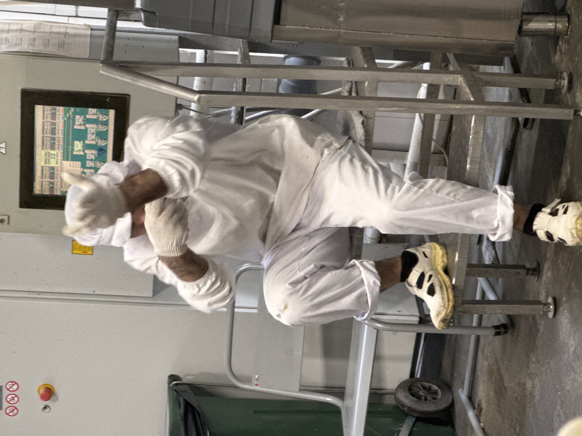 An individual wearing a white full-body protective suit, white cap, and white gloves is seated on a silver metal stool. The suit appears to be soiled in places. The individual's right hand is raised, with fingers extended in a gesture, while their left hand is in front of their face. They are wearing white shoes with yellow soles and black accents, with black socks visible. The scene appears to be an industrial setting, with machinery and control panels in the background. A green bin with a wheel is visible to the left. The floor is concrete and shows some debris. The lighting is artificial. The image does not contain any specific elements that place it in Dedovsk, Russia.