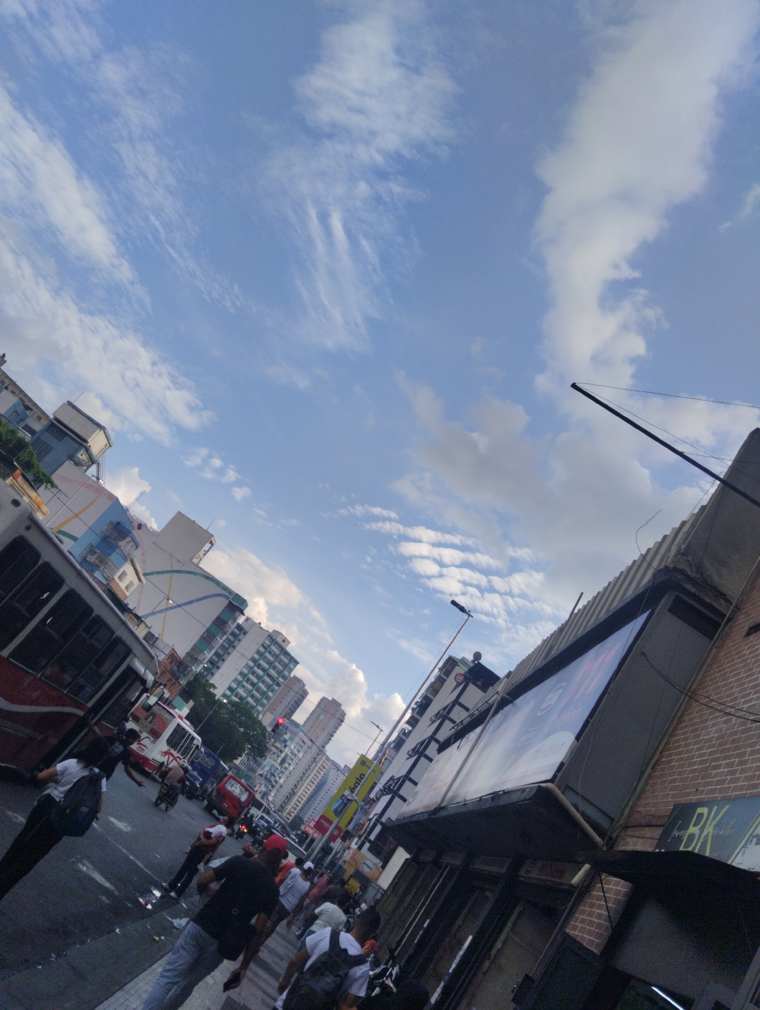 The image shows a busy street scene in an urban environment. People are walking on the sidewalks and crossing the street. Various vehicles, including buses and possibly a fire truck, are visible in the street. The architecture suggests a densely populated city, with tall buildings lining the street. The sky is blue with scattered clouds. A building with a "BK" sign is visible. The overall impression is of a bustling city street during daylight.