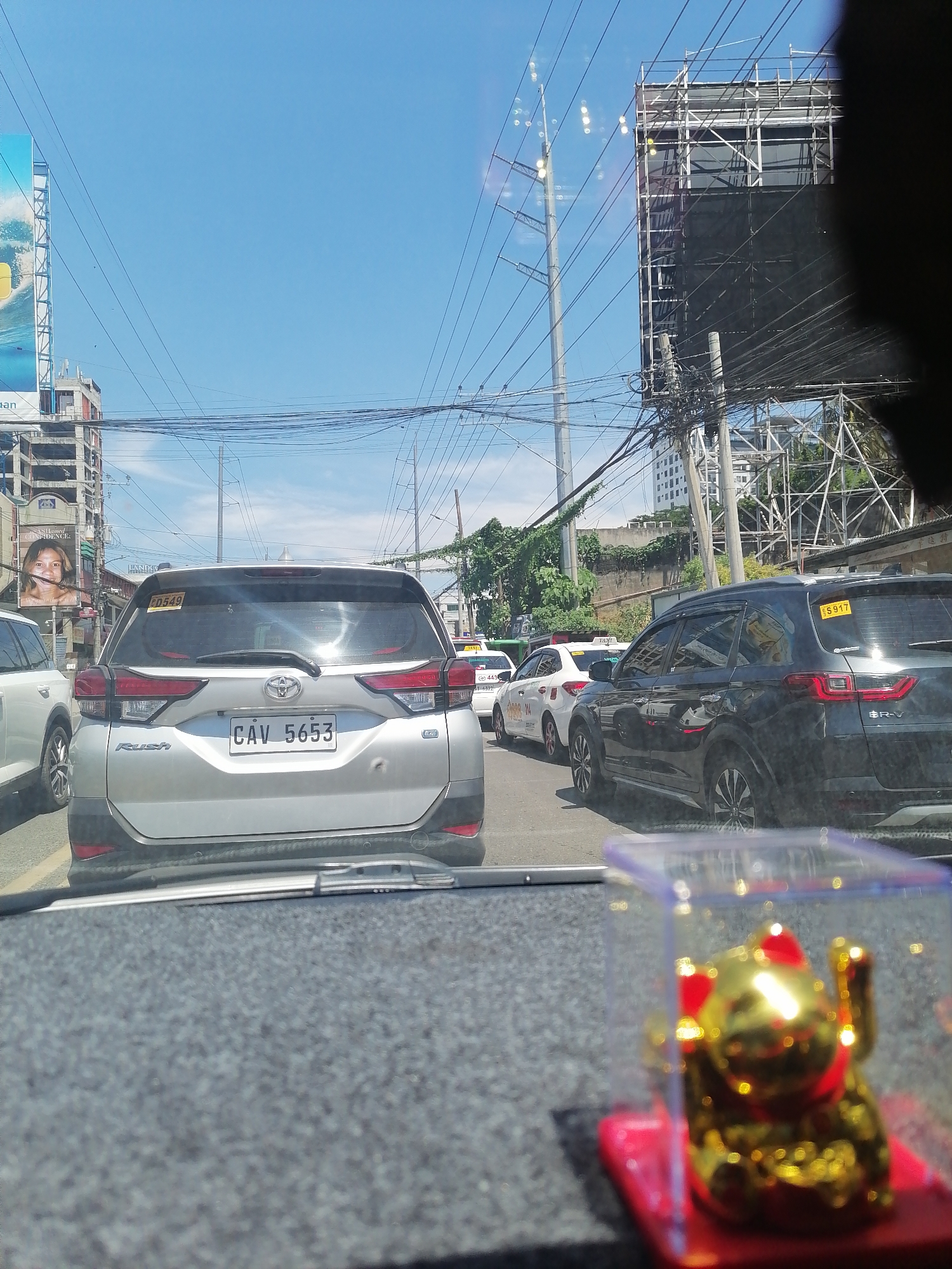 The image was taken from inside a vehicle, looking out at a street scene in Davao City, Philippines. It appears to be daytime with a clear blue sky. The foreground shows the dashboard of the vehicle, with a small golden lucky cat figurine in a clear plastic case to the right.
The midground features a silver Toyota Rush SUV with the license plate "CAV 5653". Behind it, a dark-colored Honda BR-V SUV is visible. Several other vehicles, including white taxis, are in traffic behind these main subjects. Overhead, a complex network of power lines and utility cables spans across the sky.
In the background, buildings and structures are visible. To the left, a tall building with exposed concrete framework is under construction, alongside a billboard with an advertisement featuring a woman. To the right, a large billboard structure with a black screen is present, also surrounded by scaffolding and cables. The environment suggests an urban setting with ongoing development.