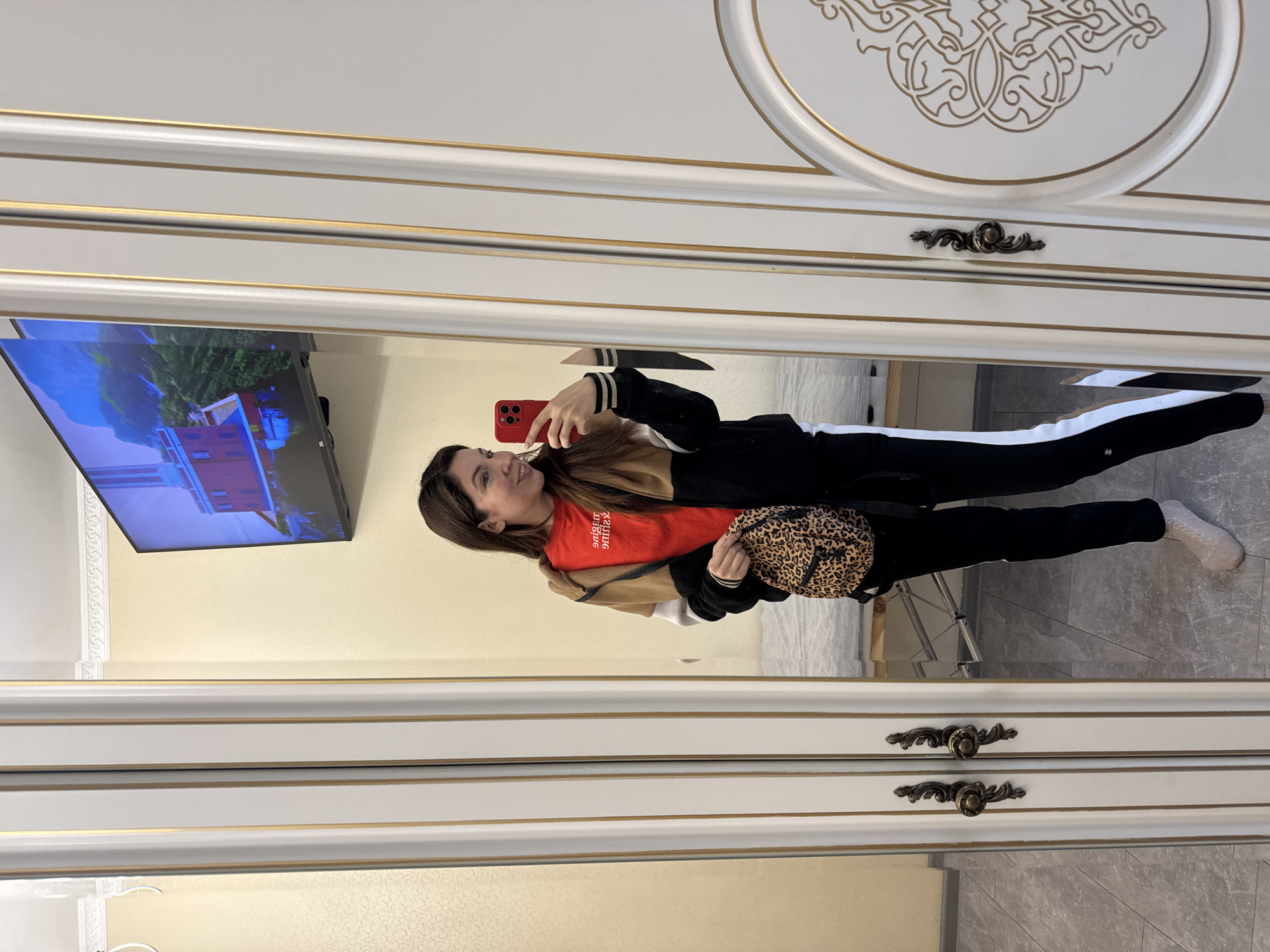 A young woman is taking a selfie in front of a large mirror. The woman is wearing a red t-shirt, a black jacket with white stripes down the arms, black pants, and white socks. She is holding a red smartphone in her right hand and a leopard-print handbag in her left hand. The mirror is framed in white with gold trim, and it reflects a television mounted on the wall, showing a building among trees, and the gray tiled floor. The reflection reveals a light-colored wall behind the woman. The location is suggested to be in the city of Kryvyi Rih, Ukraine.
