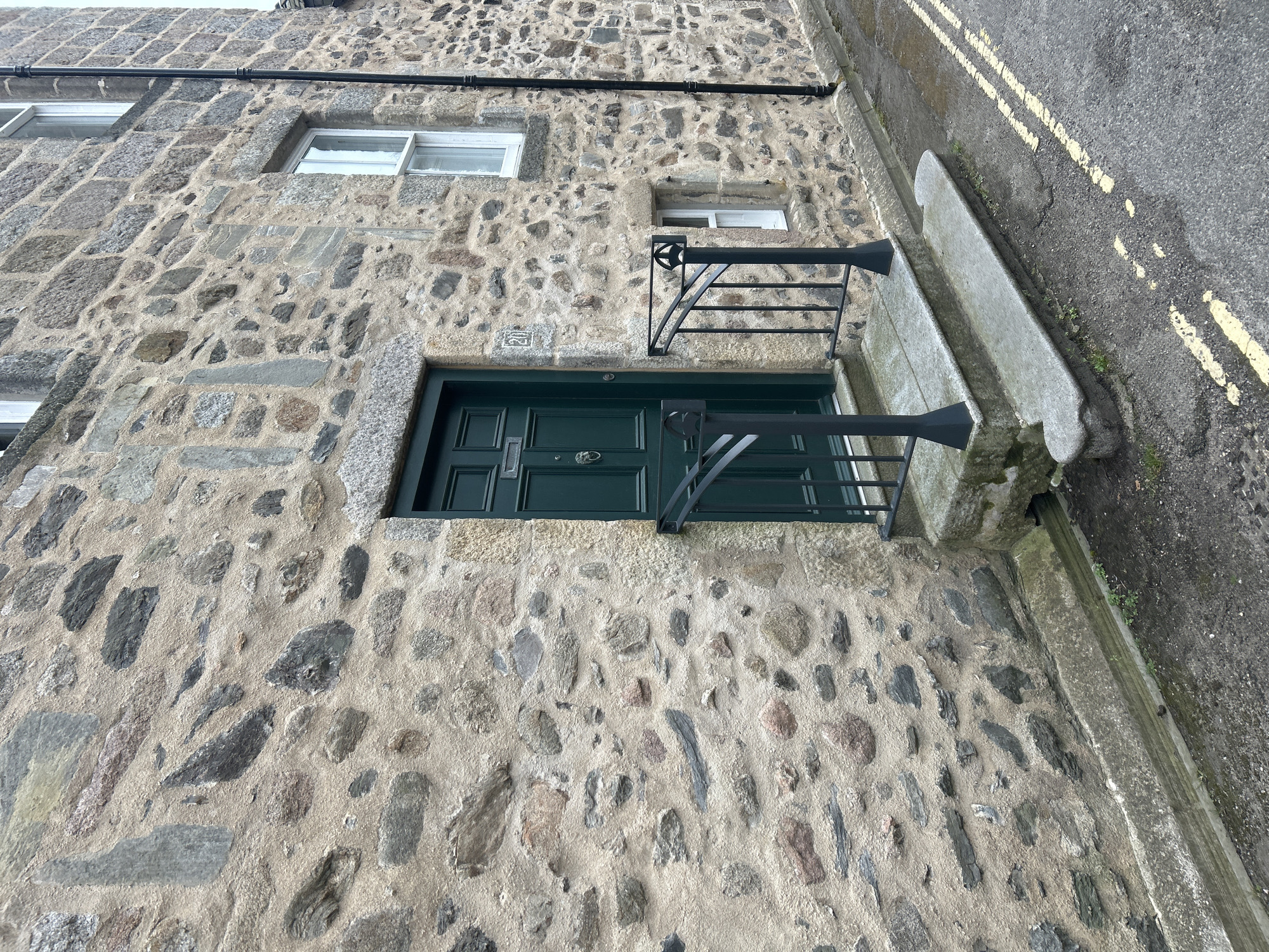 A slightly tilted, medium shot shows the exterior of a building in Aberdeen City, United Kingdom, constructed of irregularly shaped, gray, stone bricks set in mortar. The building has three windows, all with white frames and small panes, visible on the upper portion of the frame. Below the windows is a dark green, paneled door with a silver door knocker. The door is accessed by a small concrete step and flanked by black metal railings on either side. A narrow sidewalk runs along the base of the building, separated from the road by a concrete curb and a double yellow line painted on the asphalt. A black pipe runs horizontally along the upper portion of the building’s facade. Number 25 is partially visible on the wall just above the door.