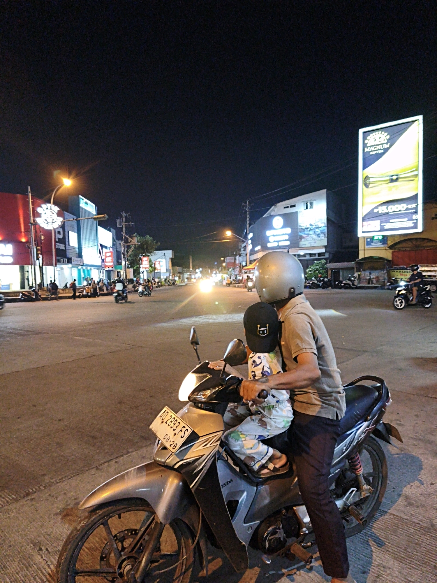 The image depicts a nighttime street scene in Surakarta, Indonesia, likely an intersection with several buildings and businesses illuminated by artificial lights. In the foreground, a person wearing a helmet is seated on a motorcycle, holding a child who is also wearing a helmet. The motorcycle's license plate reads "AD 2893 FS 08-28".
The background shows a bustling street with other motorcycles and vehicles, as well as pedestrians walking along the sidewalks. Brightly lit signs and building facades suggest commercial activity. A large billboard featuring the word "MAGNUM" is visible on the right side of the image. The street surface appears to be asphalt. The overall atmosphere is that of a lively urban environment at night.