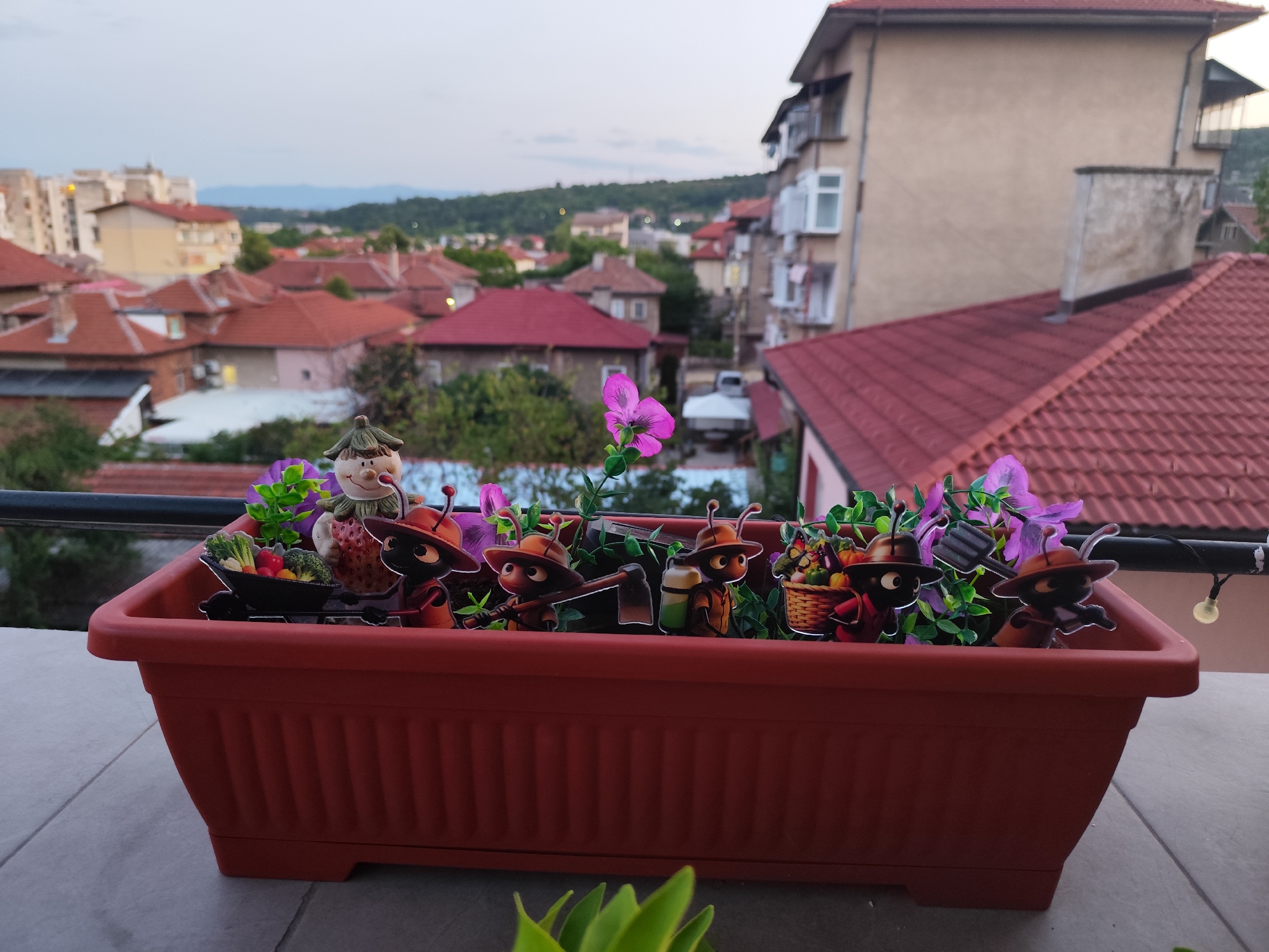 This is a photograph of a decorative planter box on a balcony overlooking a city with red-tiled roofs. The planter box is filled with artificial plants and several small figures of cartoon ants dressed in hats and carrying gardening tools and produce. In the background, residential buildings, some with balconies, are visible, along with a distant green hillside. The sky is overcast, suggesting it might be dusk or a cloudy day. The scene is likely in the city of Montana, Bulgaria, given the characteristic architecture of the buildings.