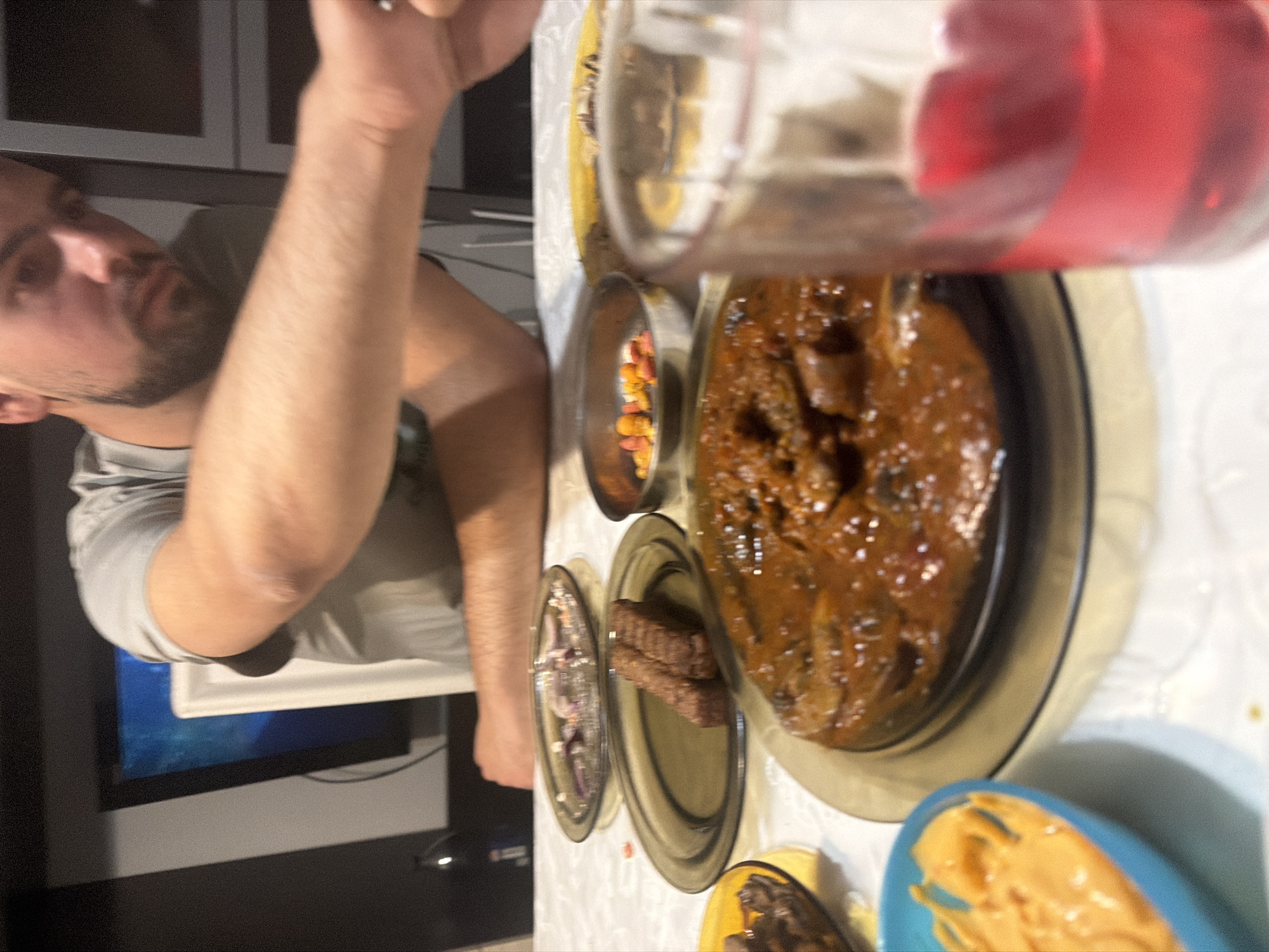 This image captures an indoor dining scene in Shumen, Bulgaria, featuring one male individual and a table laden with various food items.
In the foreground, a clear glass filled with a translucent, dark red liquid occupies the right side, out of focus. To its left, a small, bright blue bowl contains a thick, light orange, creamy substance, possibly a dip or sauce. Dominating the center-midground is a large, round, dark grey or brown glass plate holding a substantial dish. This dish appears to be a stew or goulash, characterized by a rich, reddish-brown sauce with visible chunks of meat and possibly vegetables (e.g., mushrooms) within it.
Further into the midground, behind the large stew plate, a smaller, dark grey or brown glass plate presents two elongated, rectangular, dark brown items, resembling grilled meat patties or kebabs. To the right of these, a small, polished metal bowl contains an assortment of small, colorful candies or snacks, primarily orange, yellow, and red. Behind this, another dark grey or brown glass plate is mostly empty, but shows scattered remnants of thinly sliced red onions, suggesting it previously held a salad or garnish. A partial view of a yellow plate with dark food is visible on the far left. The table is covered with a white tablecloth, showing some minor wrinkles.
An adult male, with short dark hair and a beard or stubble, is seated at the table, his upper body and arms visible. He is wearing a grey t-shirt. His right arm is extended forward, resting on or just above the table, with his hand partially obscured. His left arm is bent, resting near the edge of the table. His gaze is directed downwards towards the table.
The background consists of dark furniture or wall panels. To the left of the man, a television screen displays a glowing blue image. A white chair back is visible between the man and the screen. A dark bottle with a blue label is partially visible behind the food items on the left side of the table. The lighting is artificial, creating some shadows on the table and surrounding environment.