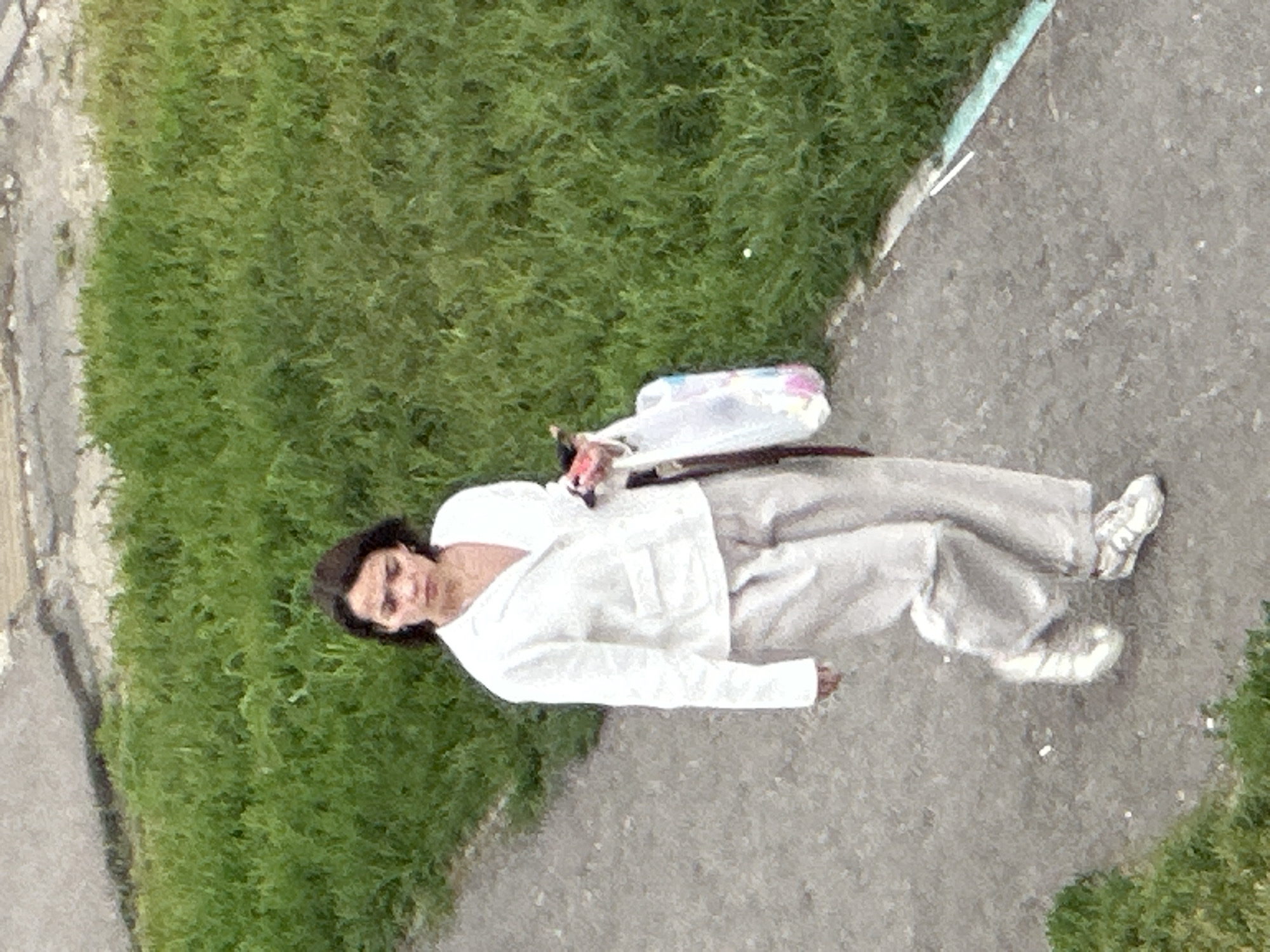 A woman walks on a paved path next to a grassy area. She is wearing a white jacket, light-colored pants, and white sneakers. She is holding a pink object in her right hand and a white plastic bag in her left hand. The image appears to be taken from a slightly elevated perspective. The environment suggests an outdoor urban or suburban setting. The presence of specific landmarks or distinctive features that would pinpoint the location in Podolsk, Russia, is not evident in the provided image.