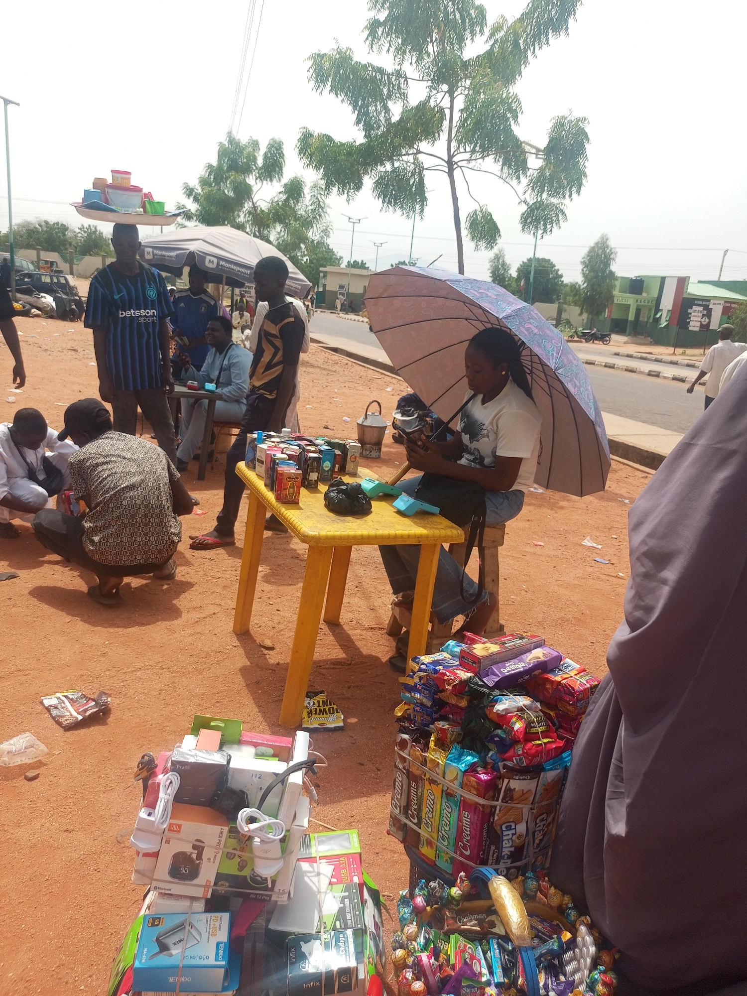 The image captures a vibrant outdoor market or street vending scene in Gombe, Nigeria, under bright daylight. The ground is unpaved and reddish-brown.
Several individuals are present, engaging in various activities. In the foreground, a woman sits on a stool under a patterned umbrella, holding a mobile device. She is seated next to a yellow plastic table laden with small boxed items, likely perfumes or cosmetics. Another man is seated opposite her, with a light blue "Moniepoint" point-of-sale device visible on the table. Two men are crouched on the ground nearby, seemingly inspecting merchandise. Further back, other people stand, including a man wearing a blue and black striped "betsson sport" shirt, and another balancing a tray of plastic containers on his head.
The scene showcases a variety of goods for sale. Next to the woman, a basket holds numerous packages of snacks and biscuits, including "Cremo," "Chak-De," and "Deemah" brands. Adjacent to this, a large collection of electronic accessories is displayed, featuring items like power banks, chargers, and headphones. Visible brands and text on these products include "JBL 90," "TENKOO," "AIR31 PRC," "Infinix," and specifications like "120W FAST CHARGE" and "2USB 3.1A."
In the background, a large tree provides some shade, and distant buildings, one painted green and white, can be seen along a paved road where a motorbike is parked. The overall atmosphere suggests a bustling commercial hub.