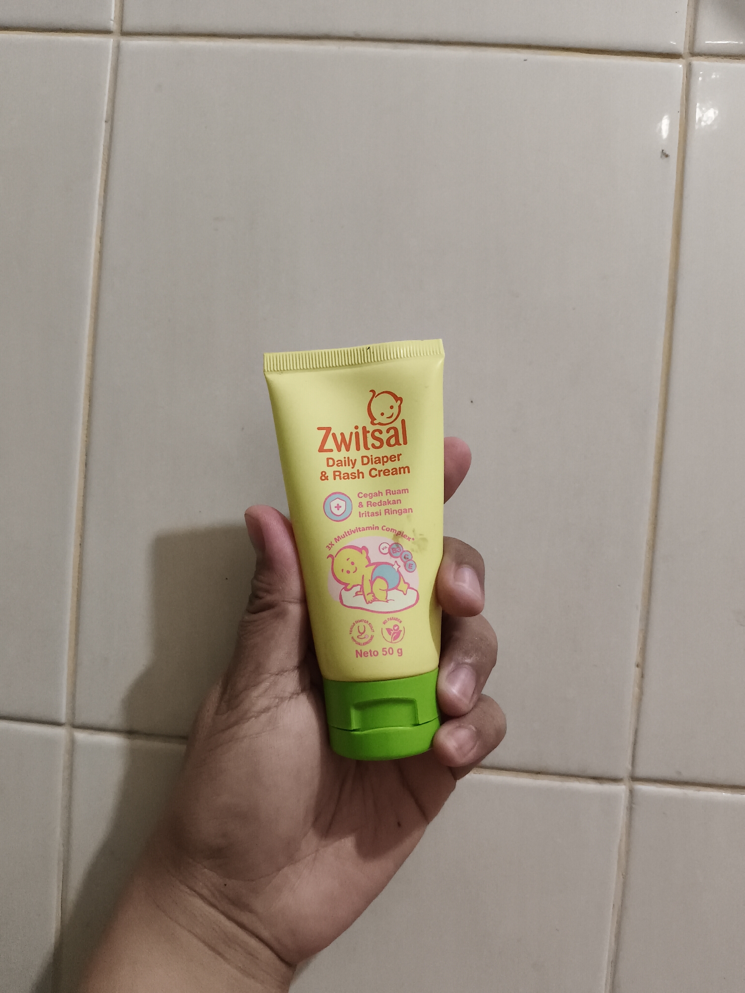 A person's hand holds a yellow tube of Zwitsal Daily Diaper & Rash Cream against a background of light-colored tiled walls. The tube features text including "Zwitsal," "Daily Diaper & Rash Cream," and "3X Multivitamin Complex," as well as an illustration of a baby. The cream tube is capped with a green lid. The tiled walls appear to be in a bathroom or similar area. The image does not contain information about the specific location within Tangerang, Indonesia.