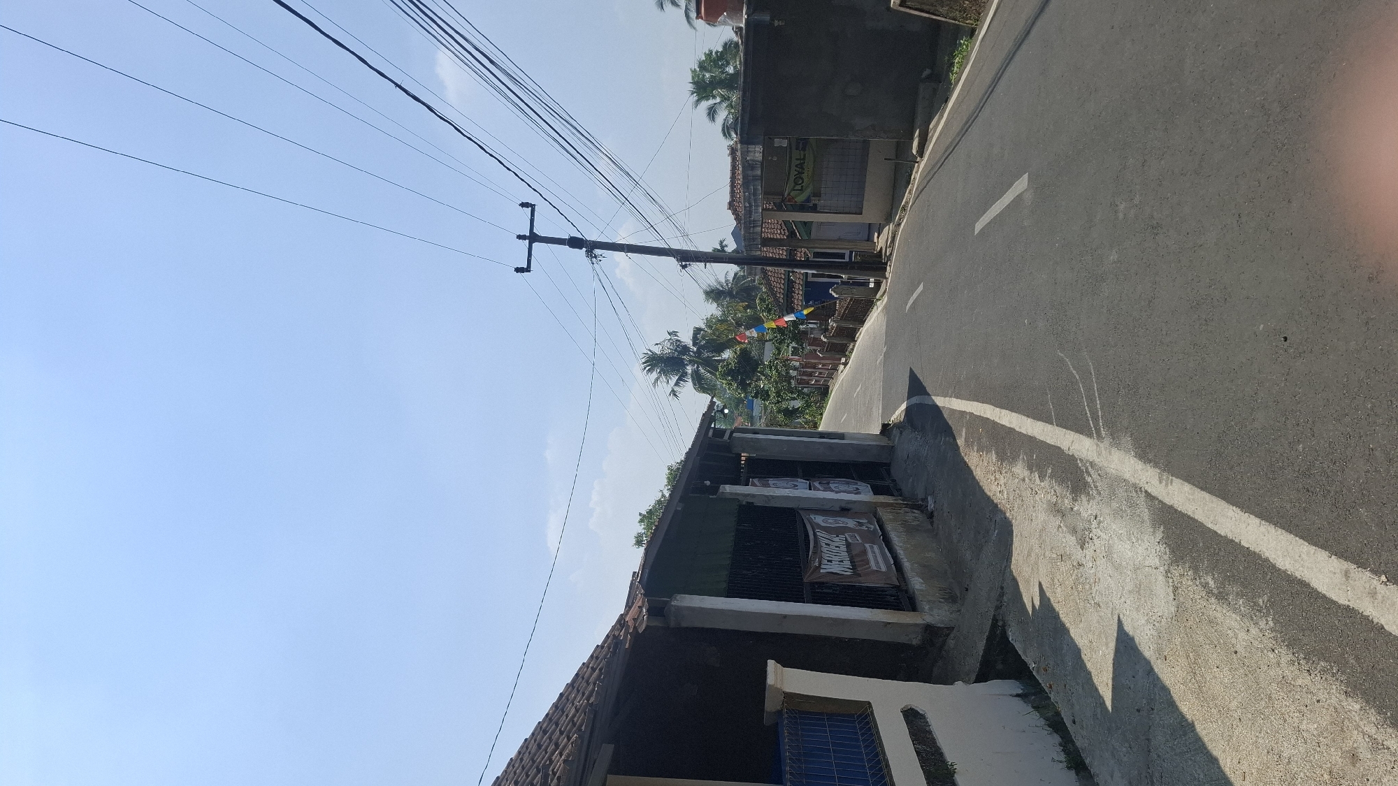 The image depicts a sunny, clear day in a rural area of Ciharashas, Indonesia, likely a village or small town, judging by the presence of residential buildings and a narrow asphalt road. The road curves to the right and ascends slightly. On the left, a low-slung building with a tiled roof and concrete pillars is visible, featuring a banner with the word "MERDEKA" (Independence) written on it, suggesting a celebration or national holiday. A blue fence is partially visible to the far left. Across the road, more residential structures are present, some with fences and tropical trees. A prominent utility pole with multiple power lines stands on the right side of the road, leaning slightly. Several small Indonesian flags are strung across the road in the mid-ground, adding to the festive atmosphere. The overall impression is of a quiet, everyday scene in a tropical village.