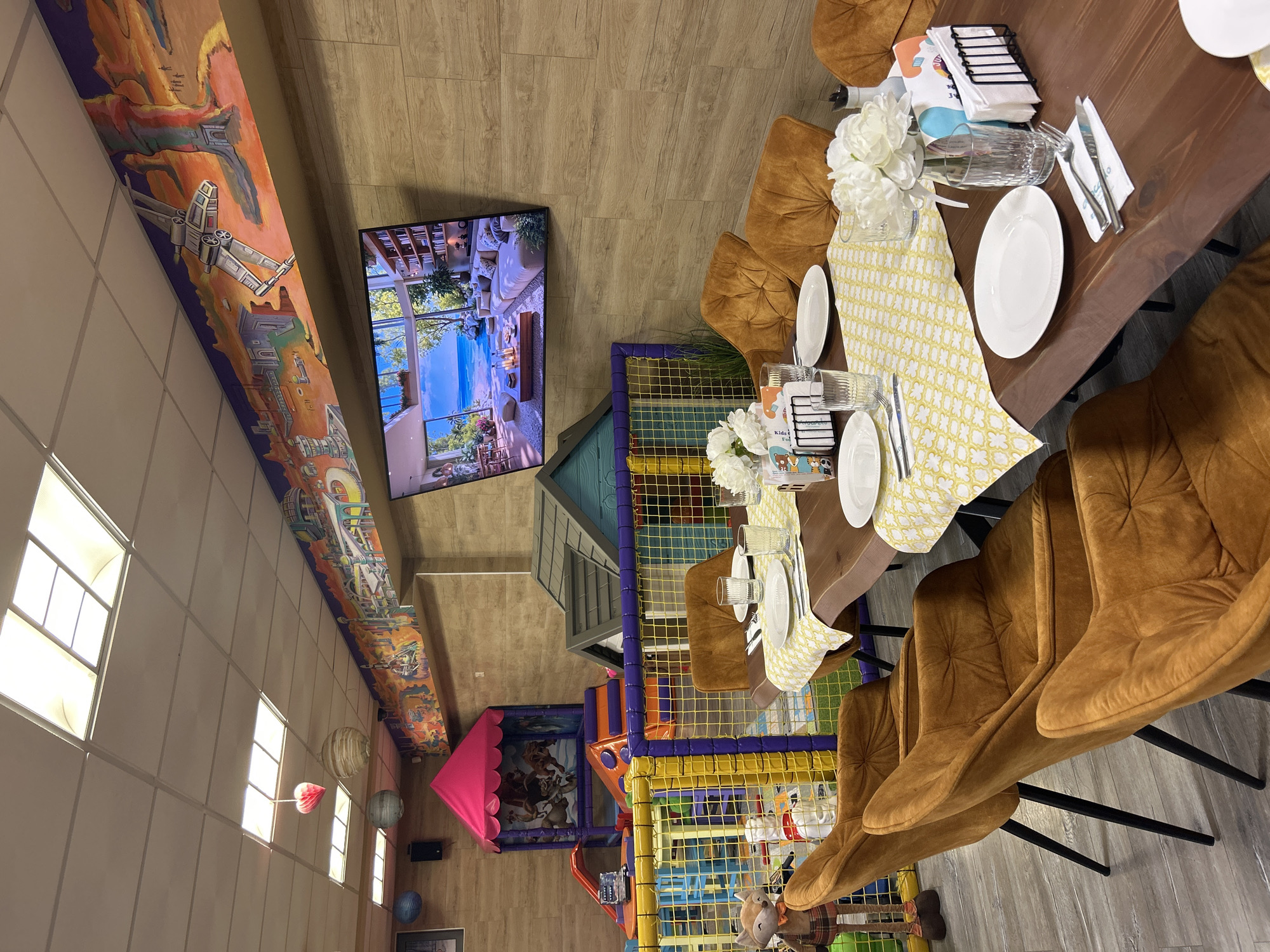 The image captures the interior of a dining area combined with a children's play space. A long wooden table is set for a meal, featuring white plates, cutlery, napkins, glasses, and floral arrangements. The table is covered with a yellow patterned runner. Brown velvet chairs surround the table. Beside the dining area, there is a multi-level playground structure with colorful plastic tubes, netting, and slides, a pink tent-like structure is also visible. A large screen is mounted on the wall displaying a beach scene. A mural adorns the upper part of the wall. The location is a restaurant or family entertainment center likely in Plovdiv, Bulgaria. There are no people visible in the image.