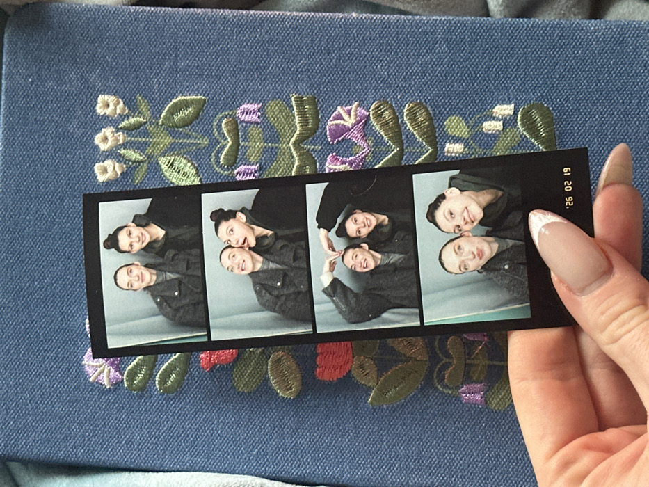 A photo strip of four images displays two individuals, seemingly a parent and child, posing playfully in each frame. The photo strip is held by a hand with manicured nails, with the date "26 02 1992" printed on the side. The backdrop features a blue textured surface adorned with floral embroidery.