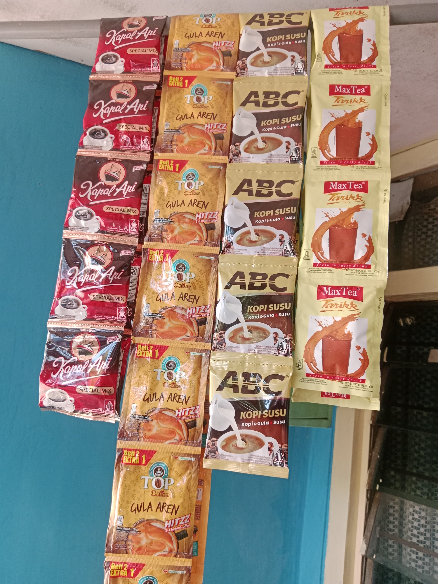 A vertical, eye-level, medium shot captures a display of assorted coffee and tea sachets hanging against a blue wall and a window with green blinds. The sachets are organized in columns, with some slightly overlapping. On the left, three rows of "Kapal Api Special Mix" sachets are visible, each featuring a red and brown design with an image of a steaming cup of coffee. To the right of these, a column of yellow "Top Coffee Gula Aren" sachets, each displaying a cup of coffee with palm sugar and text indicating "Beli 2 Extra 1." Further to the right, a column of pale yellow "MaxTea Tarikk" sachets, each illustrating a glass of iced tea with splash effects, is present. Another column of "ABC Kopi Susu" sachets, with a brown and beige design depicting a cup of coffee being poured with milk, is also visible. The lighting appears natural, suggesting an outdoor or well-lit indoor setting. The scene is static, with no visible action.