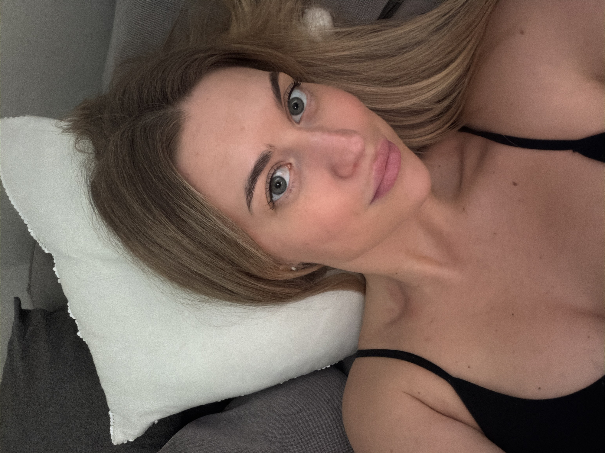 A young adult Caucasian female is depicted in a close-up, selfie-style portrait, reclining indoors. Her long, light brown hair, parted centrally, appears slightly disheveled, suggesting a relaxed posture. She has blue-grey eyes looking directly at the camera, and full, softly pink lips that are slightly parted. Her skin is clear, with scattered small moles visible on her chest and upper arm. She is wearing a black camisole or tank top, with a thin strap visible on her right shoulder. Behind her head, a white throw pillow with a decorative ruffled edge is visible against dark grey upholstered furniture. The lighting is soft and even, indicating an indoor environment. The location of this scene is Sofiivska Borshchahivka, Ukraine.