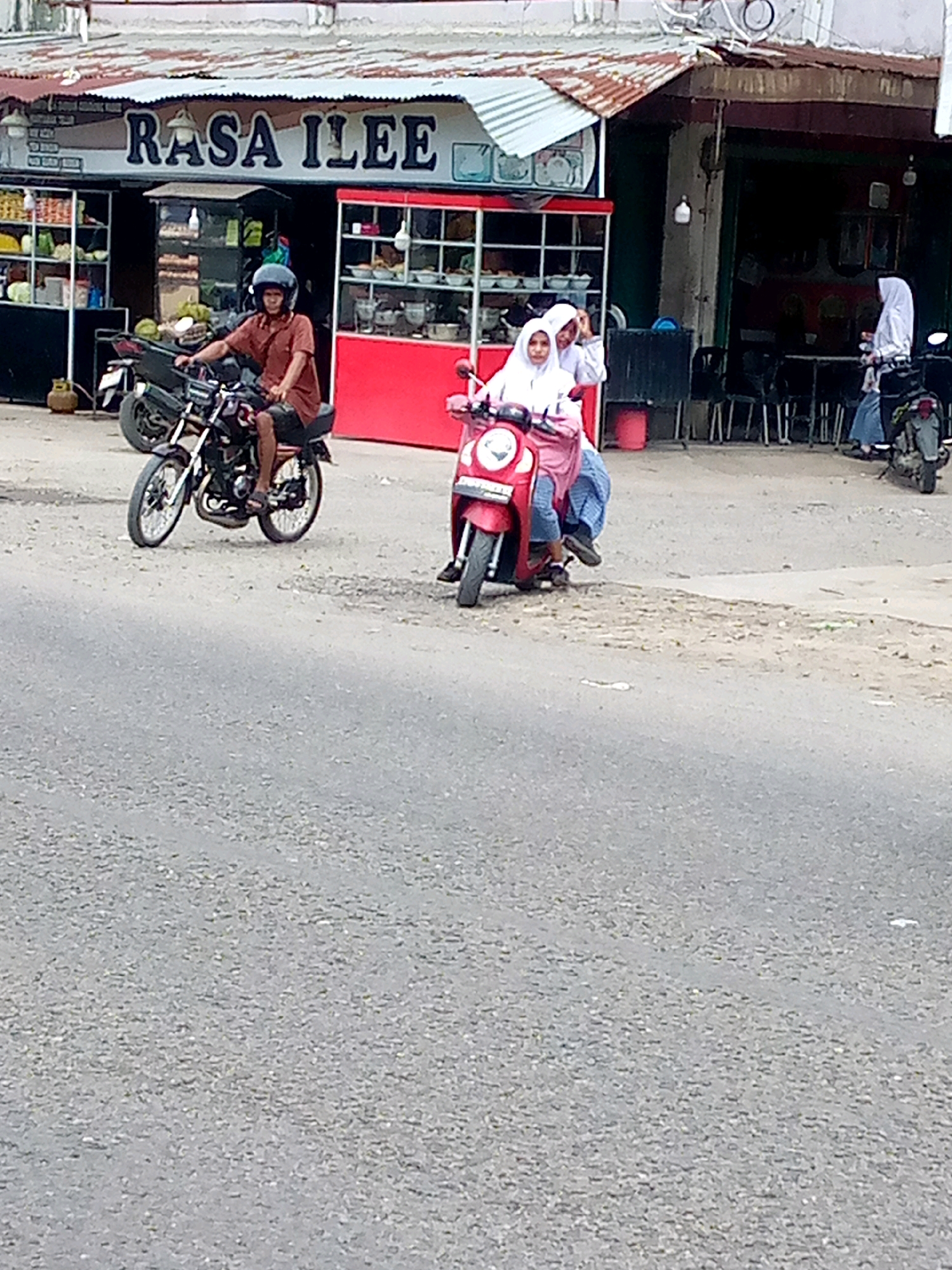 In Keudebayu, Indonesia, a man wearing a brown shirt and a dark helmet rides a black motorcycle. Two young women in white headscarves are on a red scooter, with the woman in front wearing a white hijab and the woman behind wearing a white hijab and a pink head covering. The scooter has a license plate that is partially obscured. A woman in a blue hijab and a white shirt is visible in the background, also on a motorcycle. The scene takes place in front of a store with a sign that reads "RASA ILEE" in blue lettering against a white background. The store has shelves stocked with various items and a red structure with glass doors. The ground is covered in gravel and asphalt.