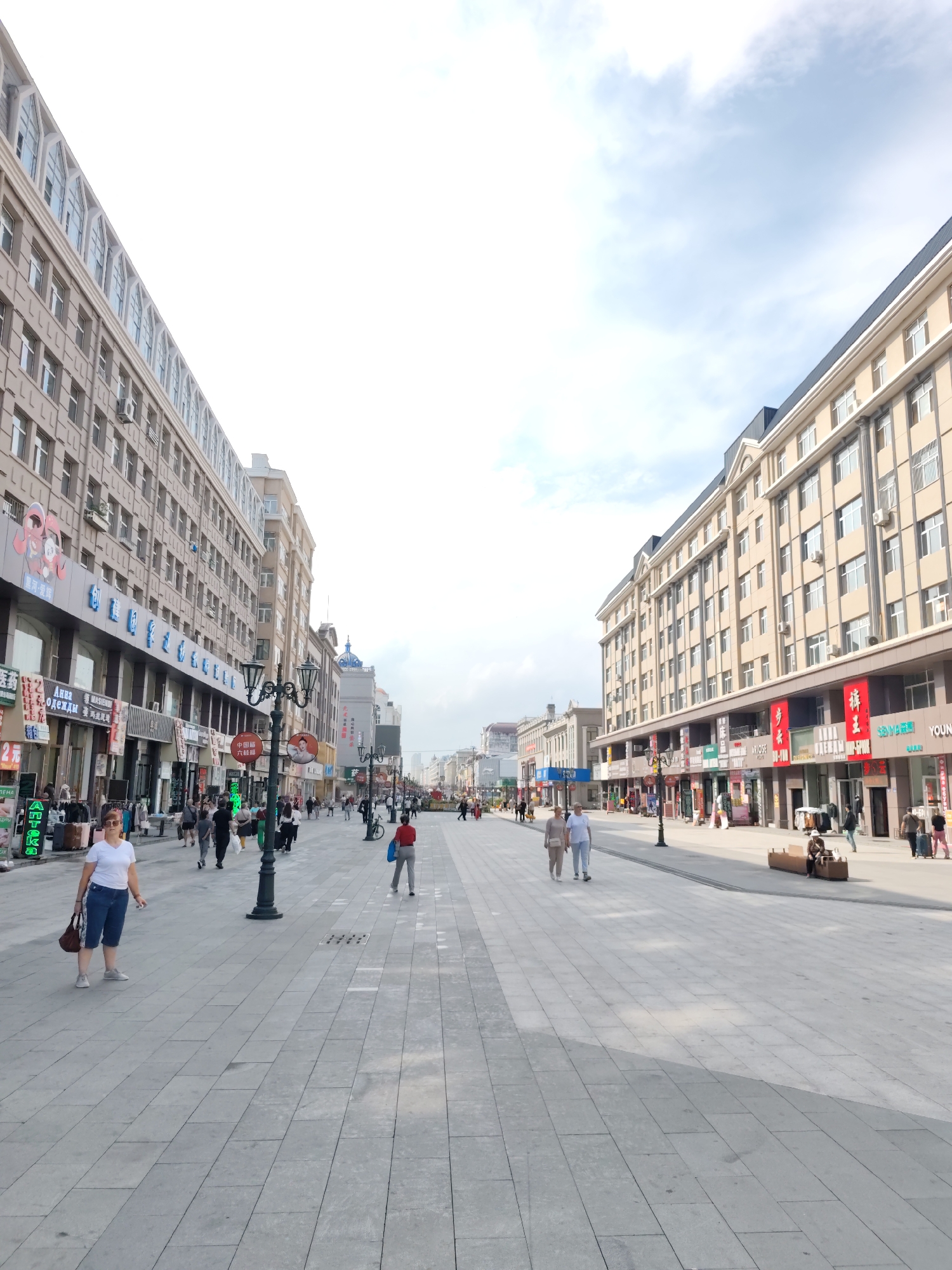This is a ground-level, eye-level, medium shot of a pedestrian street in Aihui, China, on a bright, overcast day. The street is paved with large, light gray square tiles. The view is looking down the long, wide street, flanked on both sides by multi-story buildings with a light beige facade. The buildings have numerous windows and some architectural detailing. Shops with signs in Chinese are visible at street level.
On the left side of the street, a woman in a white t-shirt and blue shorts stands looking towards the camera, holding a brown purse. She is wearing sunglasses and white sneakers. Several other pedestrians are walking in the distance. On the right side of the street, two older individuals, a man and a woman, are walking away from the camera. Further down the street on the right, several people are walking, some with luggage, and a few are seated on a modern wooden bench. Streetlights with an ornate design are positioned along the walkway. The sky is bright with some clouds.