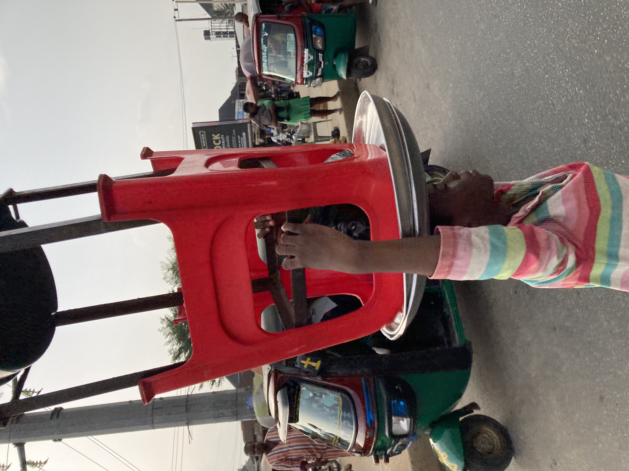 A vendor in Ura Nta Umuarandu, Nigeria, transports red plastic chairs atop a three-wheeled vehicle. He wears a striped shirt. Other people, vehicles, and structures are in the background.