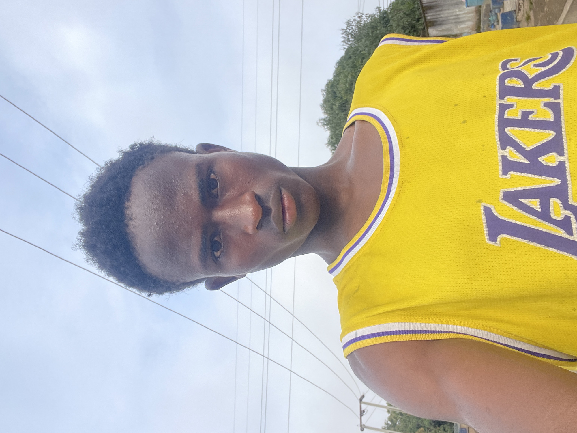 A portrait of a young Black male is captured from the chest up, facing the camera directly. He has short, dark, curly hair and is wearing a bright yellow basketball jersey with purple and white trim. The jersey has "LAKERS" written in large purple letters across the chest. His skin appears slightly shiny, suggesting sweat or humidity.
The background is an outdoor urban scene. Overhead, thick electrical wires crisscross against a pale, overcast sky. To the right of the subject, a weathered concrete wall and a large green tree are visible. The ground appears to be dirt or gravel, with some blue containers or barrels and hints of other structures in the distance, suggesting a residential or commercial area. The overall lighting is flat, typical of an overcast day. The location is identified as being in the city of Jos, Nigeria.