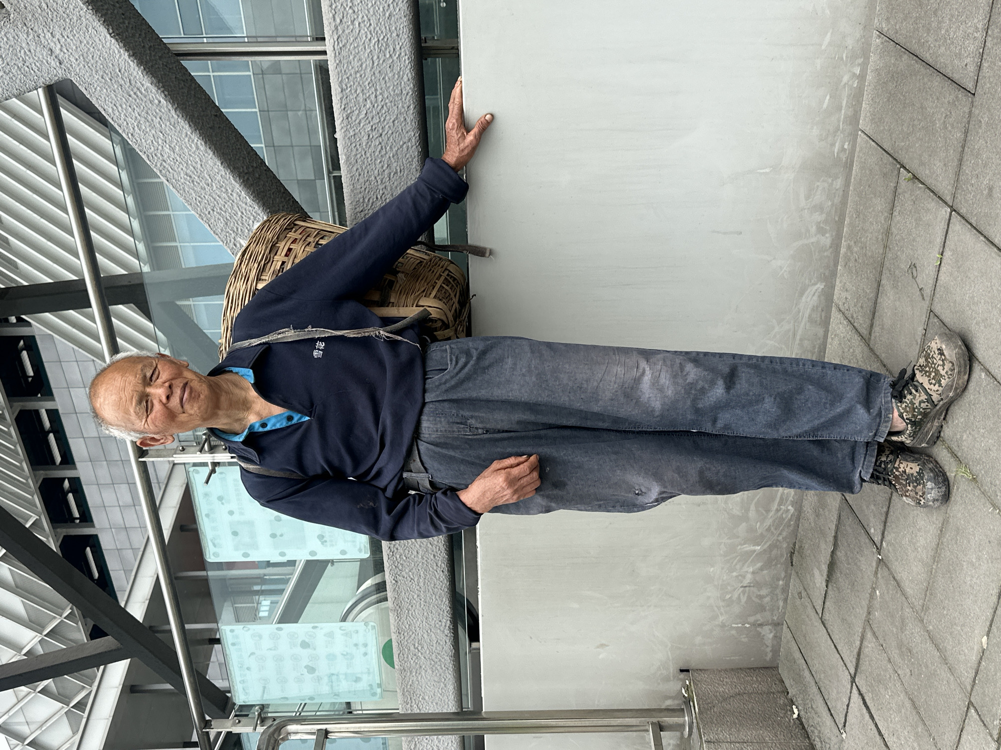 The image shows an elderly man in Jiangjin, China, wearing a dark blue long-sleeved shirt with a light blue collar, gray jeans, and camouflage-patterned sneakers. He has a woven basket strapped to his back. The man is standing on a gray tiled sidewalk, leaning against a light gray wall. In the background, there's a modern building with glass windows and metal beams. A metal railing is visible at the bottom of the frame.