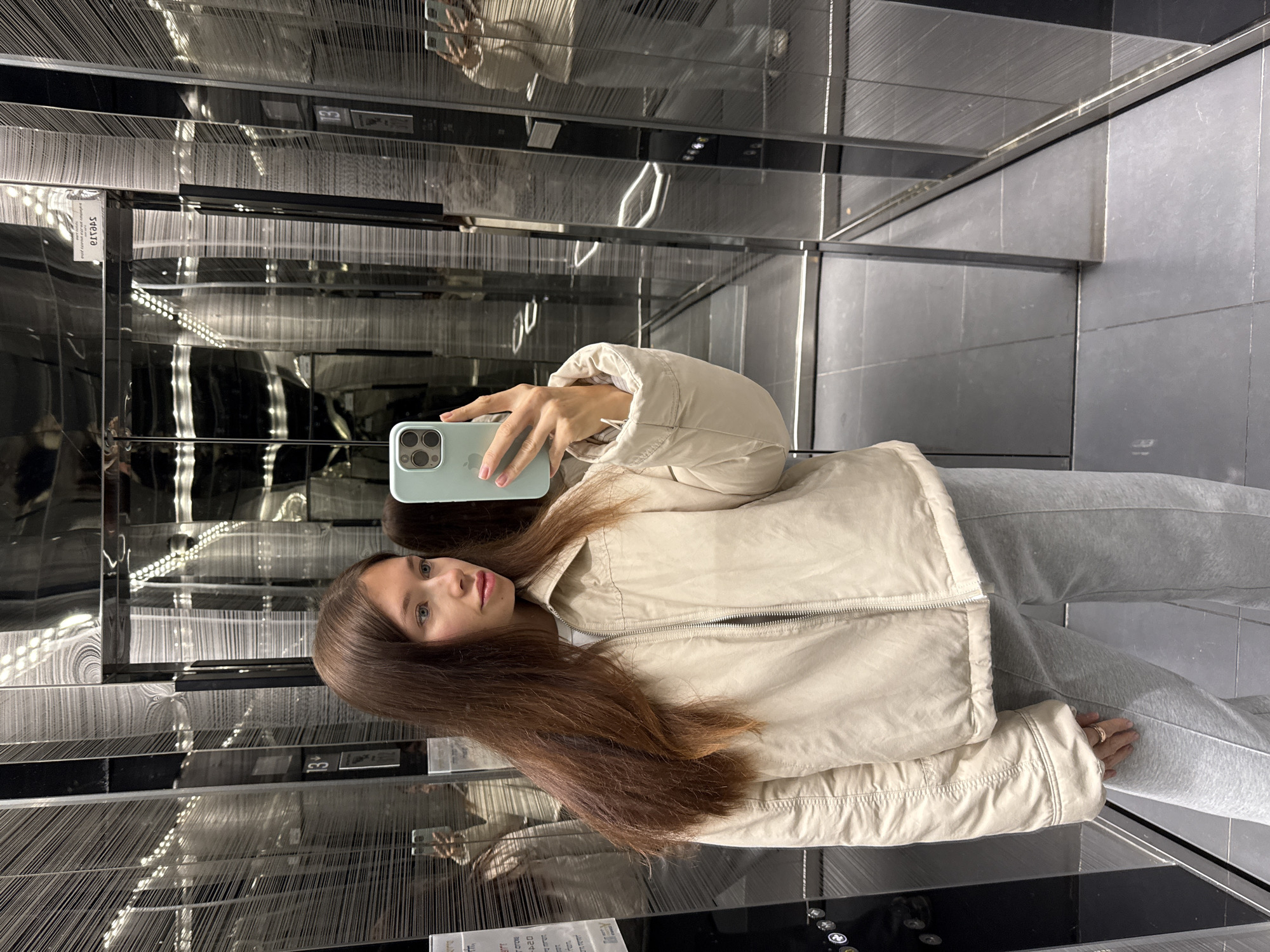 A young woman is taking a selfie in an elevator. She is wearing a beige jacket and gray sweatpants and holding a light blue smartphone. The elevator has mirrored walls with decorative lighting and a floor indicator showing "13". The elevator is likely located in a modern building in Tel-Aviv, Israel.