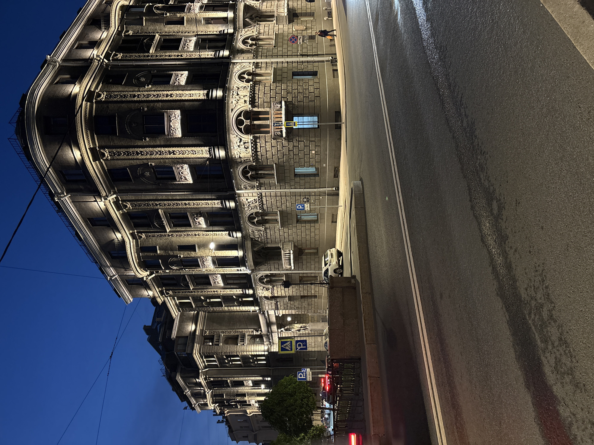 A nighttime street scene in Saint Petersburg, Russia, features a prominent building on the left side of the frame. The building's architectural style is ornate, with multiple stories and numerous windows. The building's facade is illuminated with warm-toned lights, accentuating the decorative details. To the right of the building, a street runs alongside the structure. The road surface is wet, possibly from rain, and reflects the surrounding lights. A single pedestrian walks along the sidewalk. A white car is parked along the curb. Parking signs are visible along the street. The sky is a deep blue, indicating the time of day is dusk or night. Overhead wires cross the sky. A tree is in the bottom center of the image. The overall perspective of the image is tilted to the side.