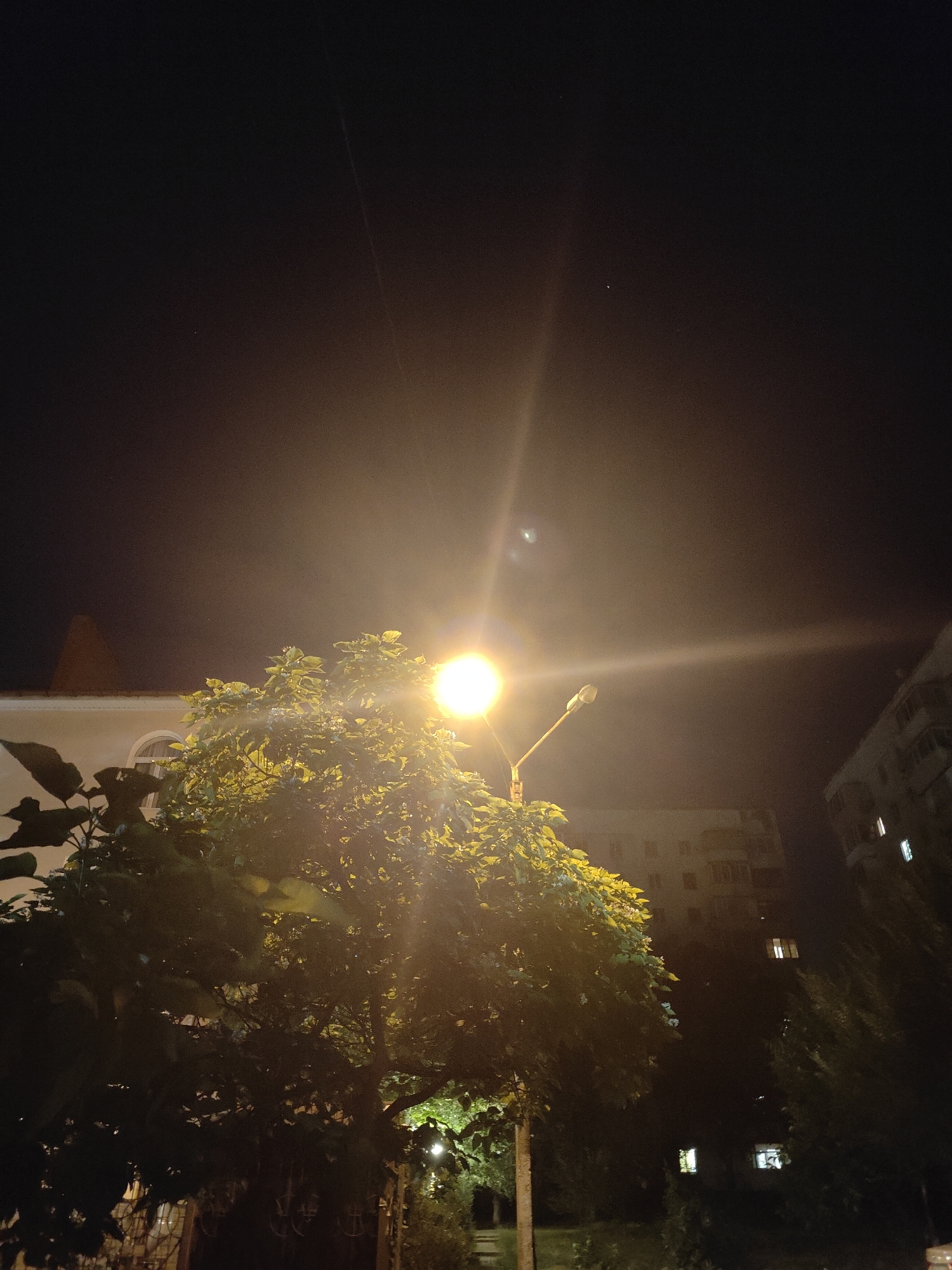 This is a nighttime, low-angle, vertical photograph taken in the city of Chornomorsk, Ukraine. A bright street lamp illuminates a leafy tree in the foreground, with its light creating a lens flare effect. Behind the tree, a building with arched windows is visible on the left, and apartment buildings with illuminated windows are seen in the background on the right. The sky is dark, with a few faint stars and a thin, faint streak of light, possibly from a passing aircraft or satellite. The overall scene depicts a typical urban night view.