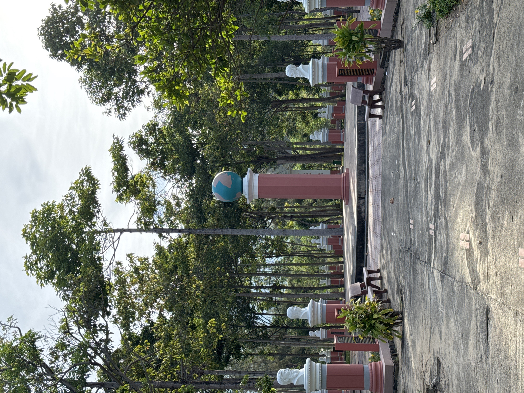 The image depicts the memorial site in South Quy Nhon Ward, Vietnam. The scene is an outdoor park-like area with a paved ground, lined with trees. In the center of the image, a large, red-colored pedestal supports a globe, symbolizing the Earth. Around this central monument, numerous smaller red pedestals are arranged in rows, each topped with a white bust of a person. Some of these busts are clearly visible, featuring distinct facial features and headwear. The busts are positioned on white, circular bases. Benches made of dark brown material are placed on the paved ground in front of some of the pedestals, and a small green plant is located next to one of the benches. The background is dominated by tall, slender trees with lush green foliage, creating a natural and somewhat somber atmosphere. The sky above is partly cloudy. The overall impression is that of a memorial or historical site dedicated to significant figures.