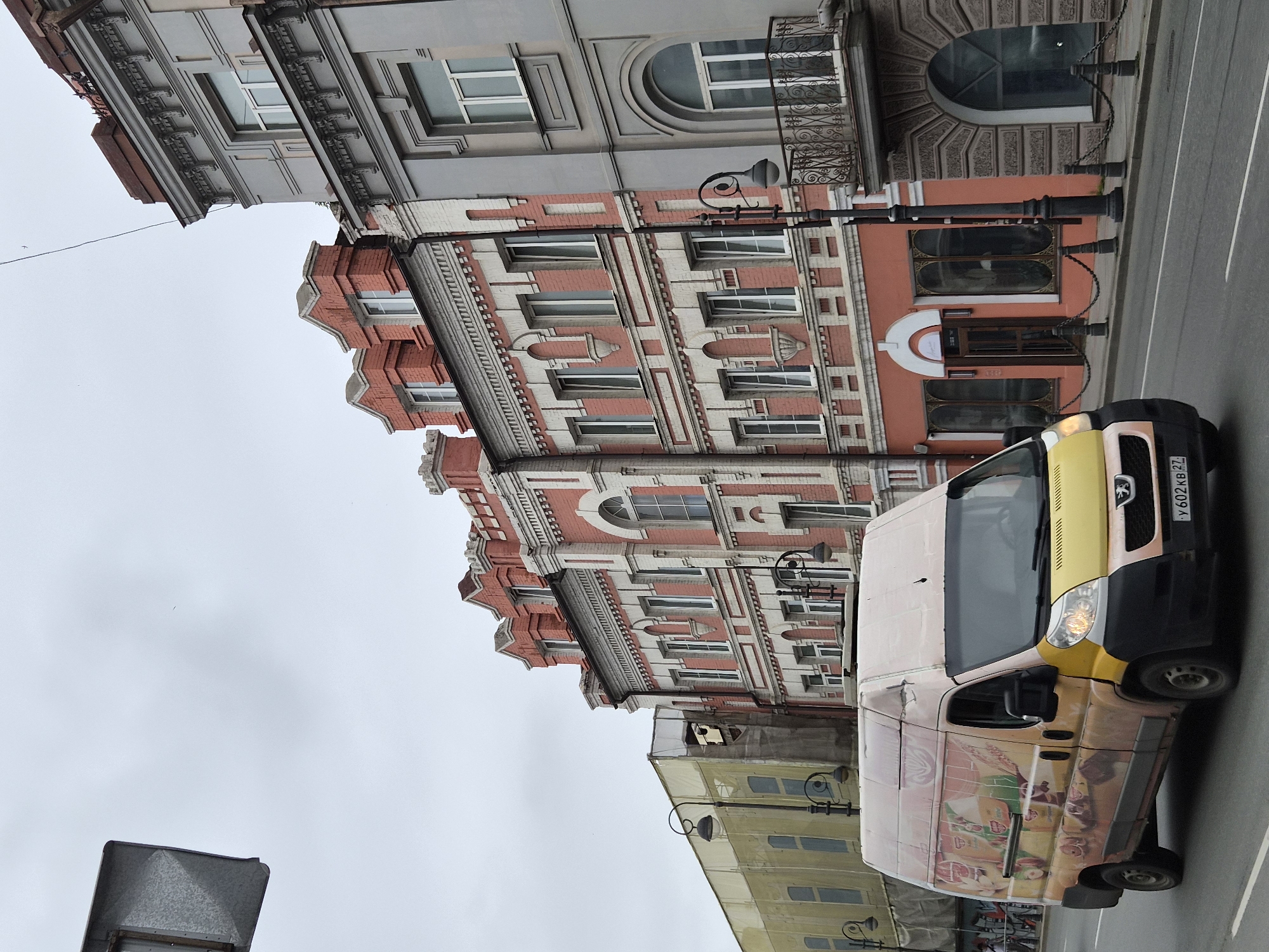 A forward-facing view of a street in Vladivostok, Russia, features a white and yellow delivery van with orange and yellow advertisements on its side, driving past ornate, multi-story buildings with red brick and grey stucco facades. The buildings have numerous windows, decorative architectural details, and balconies. A pedestrian walkway with black bollards and chains runs in front of the buildings. The sky is overcast. The street is paved with asphalt and marked with white lines. The van's license plate is visible.
