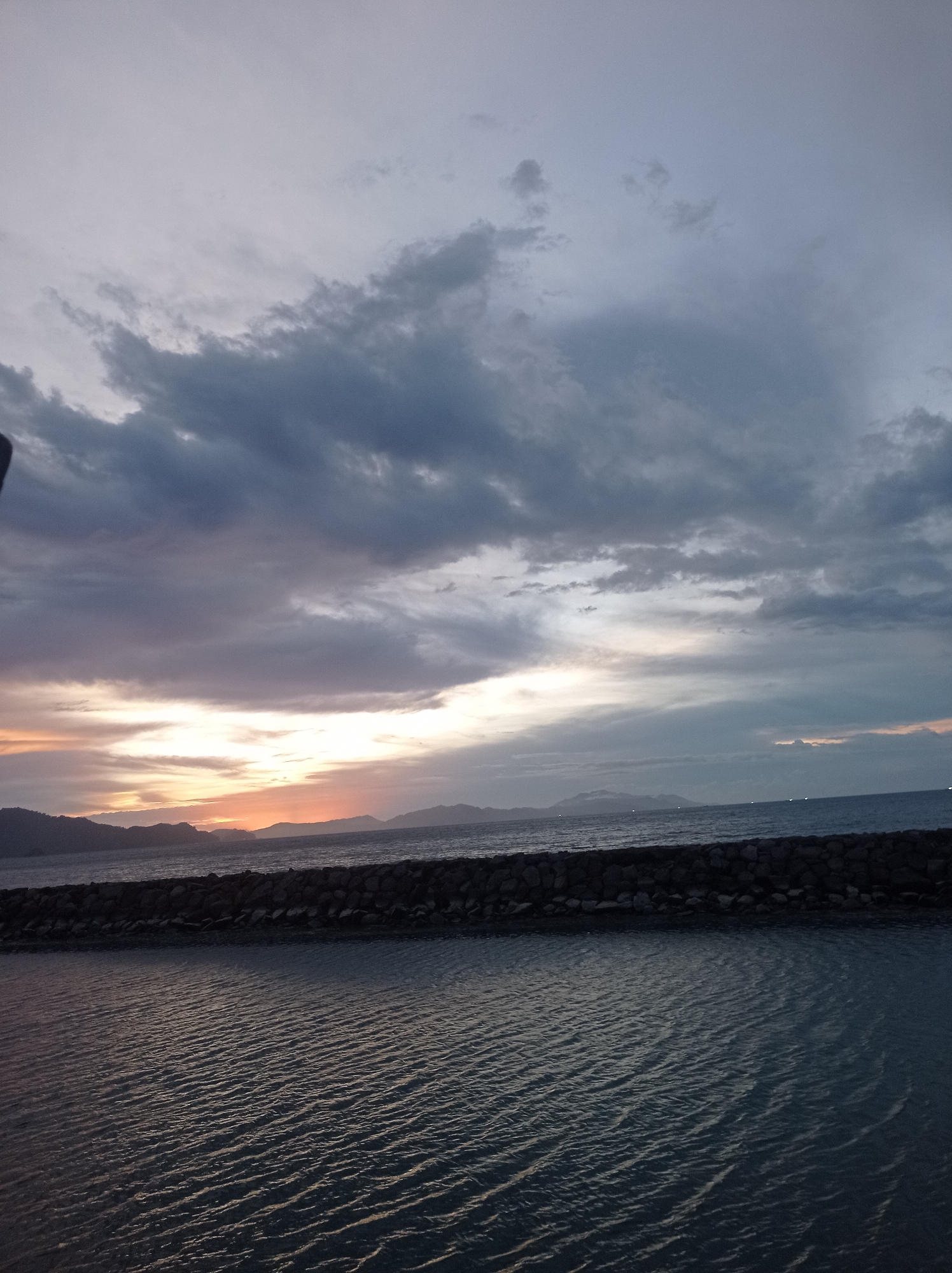 The image depicts a serene seascape at dusk, likely in Banda Aceh, Indonesia. The sky is a canvas of dramatic clouds, ranging from deep grays to softer purples and oranges, indicative of the setting sun. A band of warm light breaks through the clouds near the horizon, illuminating the distant, silhouetted mountains. In the foreground, the calm water reflects the subdued light, with subtle ripples disturbing its surface. A man-made structure, possibly a breakwater or seawall constructed from dark stones, stretches across the mid-ground, separating the inner water from the open sea. Beyond the breakwater, the sea stretches towards the horizon, where the faint lights of distant boats can be observed. The overall atmosphere is tranquil and contemplative, capturing the quiet beauty of the coastal twilight.