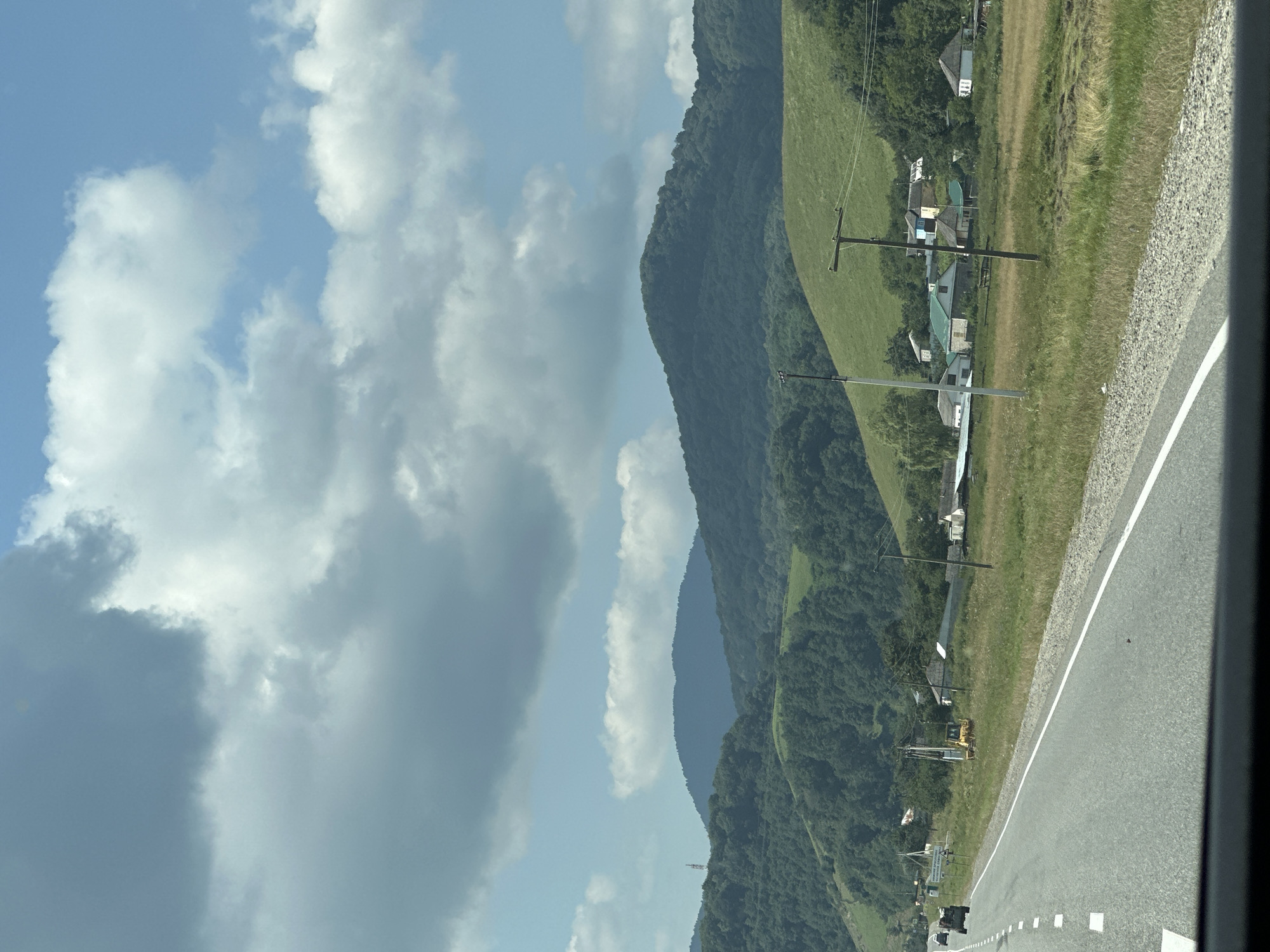 The image shows a view from a car driving along a paved road in a rural area with rolling green hills and mountains in the background. The sky is partly cloudy with a mix of gray and white clouds against a blue sky. Along the right side of the road, there is a field with dry grass and a few houses with light-colored walls and some with darker roofs. There are also utility poles and wires visible. In the distance, on a grassy slope, there is a yellow bulldozer. Further back, dense green forests cover the hills, and more distant mountains are visible. The road has white dashed lines marking the lanes and a white solid line on the right edge. Two vehicles are visible driving on the road in the distance. The scene suggests a natural landscape with scattered human settlements.