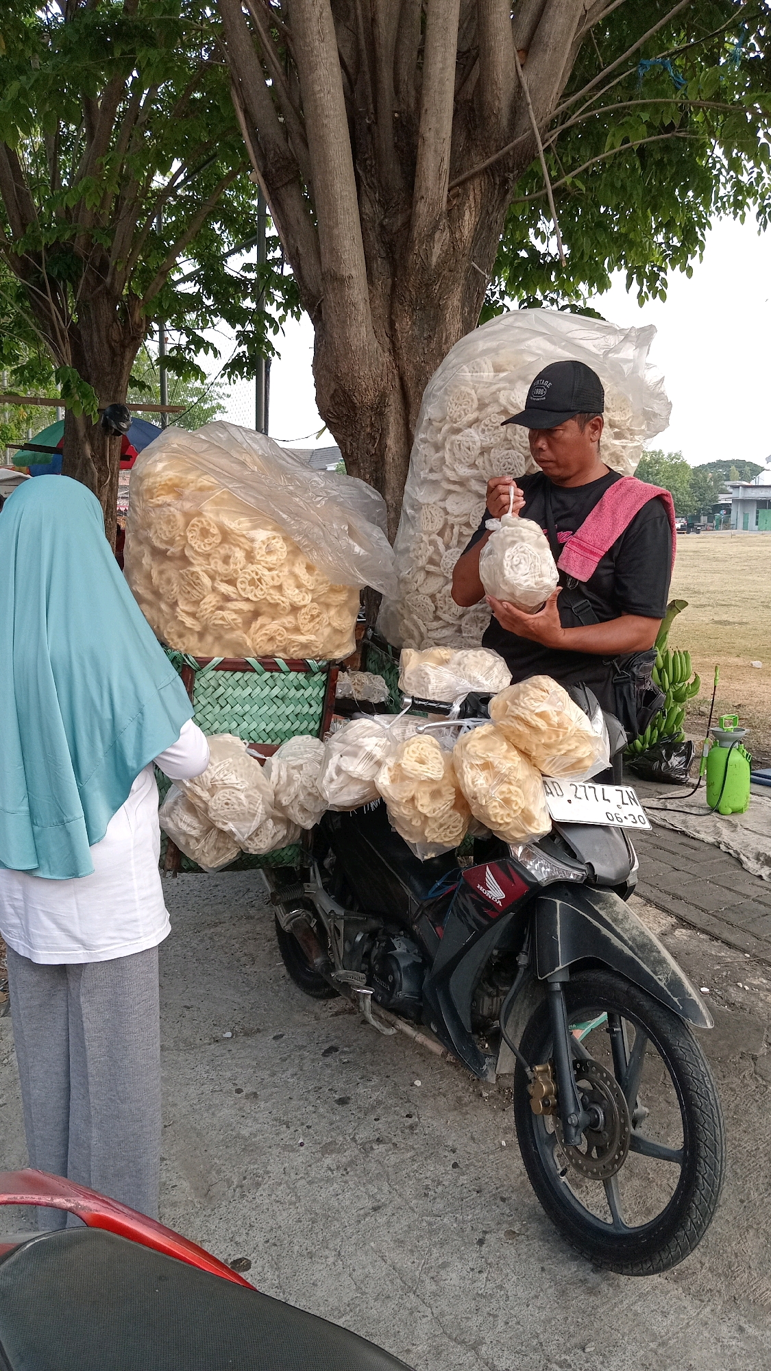 In Pongangan, Indonesia, a man wearing a black cap and a black t-shirt with a pink cloth draped over his shoulder stands beside a parked motorcycle laden with large bags of crackers. He holds a small bag of crackers in his hands, examining it. To his left, a person wearing a light blue hijab and a white shirt stands with their back to the camera, facing the crackers. The motorcycle, a black Honda, has a license plate that reads "AD 2774 ZA". Several plastic bags filled with crackers are attached to the motorcycle. In the background, large trees with textured bark dominate the scene, with foliage overhead. A green spray bottle and a bunch of bananas are visible to the right of the motorcycle. The ground is made of rough concrete. The overall setting suggests an outdoor market or roadside stall.