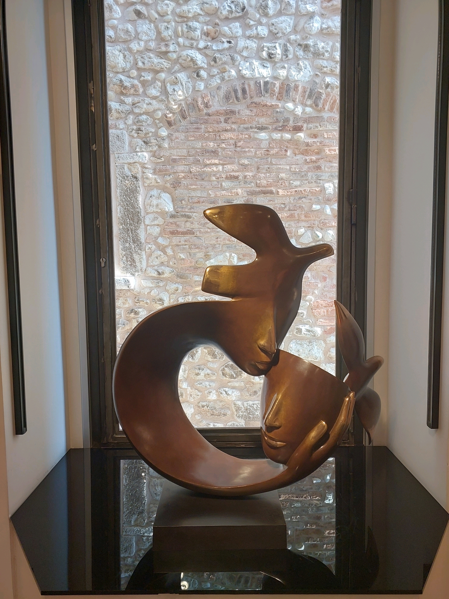The image shows a bronze sculpture of two faces intertwined with bird-like forms. The sculpture is placed on a black reflective surface in front of a window with a stone and brick wall visible through it. This scene is located in the Fondation Maeght museum in Saint-Paul-de-Vence, France. The sculpture is illuminated, and its metallic surface reflects light. The background wall has a rough texture due to the stones and bricks. The overall impression is one of artistic display within a historical setting.