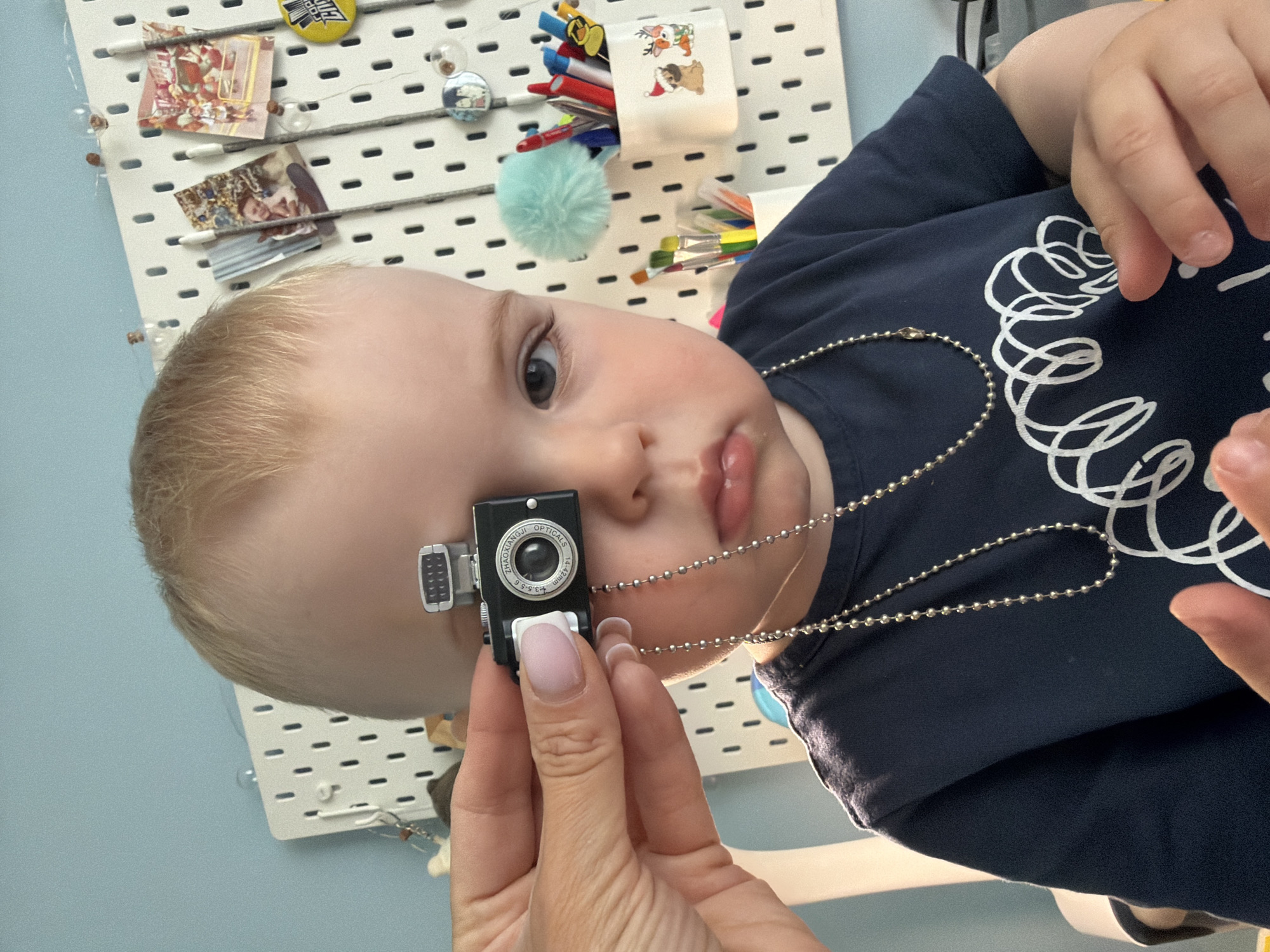 A close-up, rotated image captures a young child's face in a playful indoor setting in Votkinsk, Russia. The child, a baby or toddler with fair skin and light blond hair, looks directly at the viewer with a curious, neutral expression. An adult's hand, showing manicured nails, holds a miniature black camera-shaped toy near the child's left eye. The camera toy features "ZHAOXIANQJI OPTICALS" and "f=4.2mm" text on its lens, along with "3.5-5.6" markings, and a small silver "flash" attachment. A silver ball chain hangs around the child's neck, connected to the miniature camera.
The child wears a dark blue t-shirt adorned with white swirling patterns. In the background, a white pegboard system is visible against a light blue wall. Various items are attached to the pegboard: small decorative framed pictures, a yellow button or badge with partial text "FOR C..." and possibly "CID", a clear spherical ornament with a figure inside, and small containers holding pens/markers and paintbrushes. A light blue fluffy pom-pom is also attached to the pegboard. The scene suggests a child's room or play area, characterized by its decorative and organized elements. The activity appears to be a gentle interaction or play involving the toy camera.