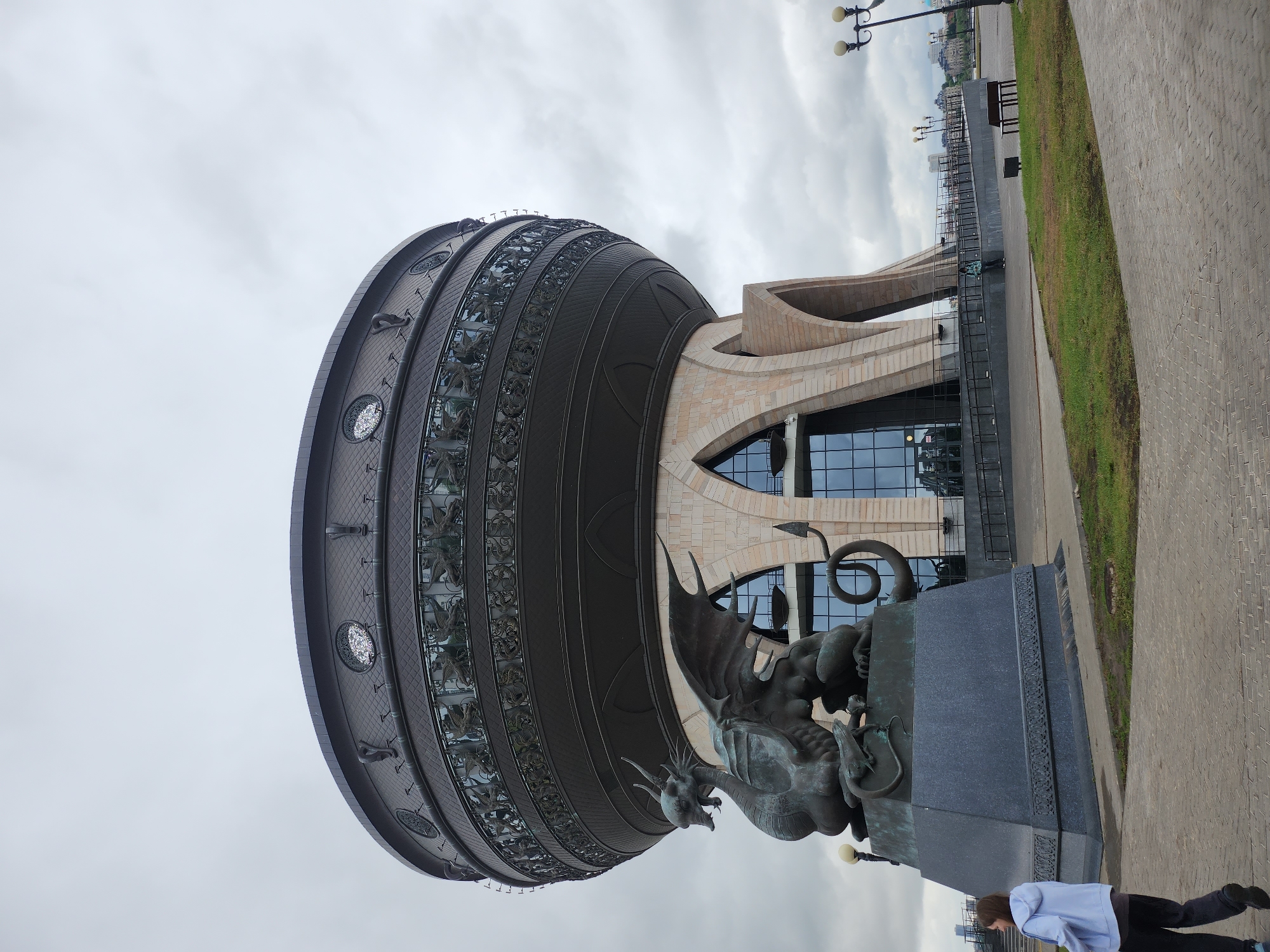 Here is a description of the image:
The image shows a large, globe-like structure with decorative elements and a statue of a dragon in front of it. The building behind the globe has a modern architectural style with large glass windows. The scene takes place on a paved area next to a patch of grass. A person in a light blue shirt and dark pants is walking along the edge of the paved area. The sky is overcast.
The location is the Wedding Palace in Kazan, Russia.