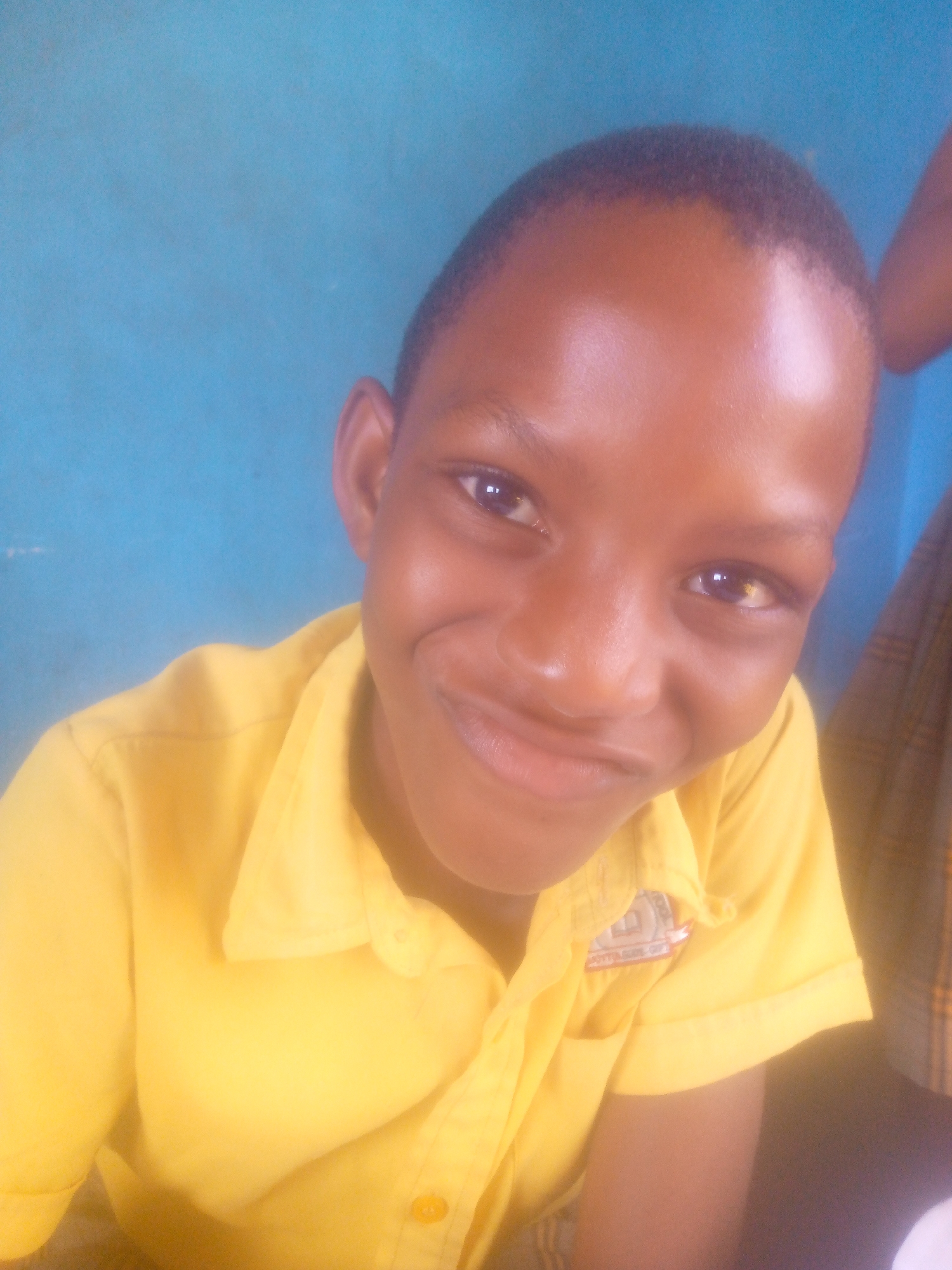 A close-up shot depicts a young, dark-skinned person, likely a child, wearing a yellow collared shirt, possibly a uniform. The background is a light blue color. The subject has a slight smile and is looking directly at the camera. The location and city are unknown.
