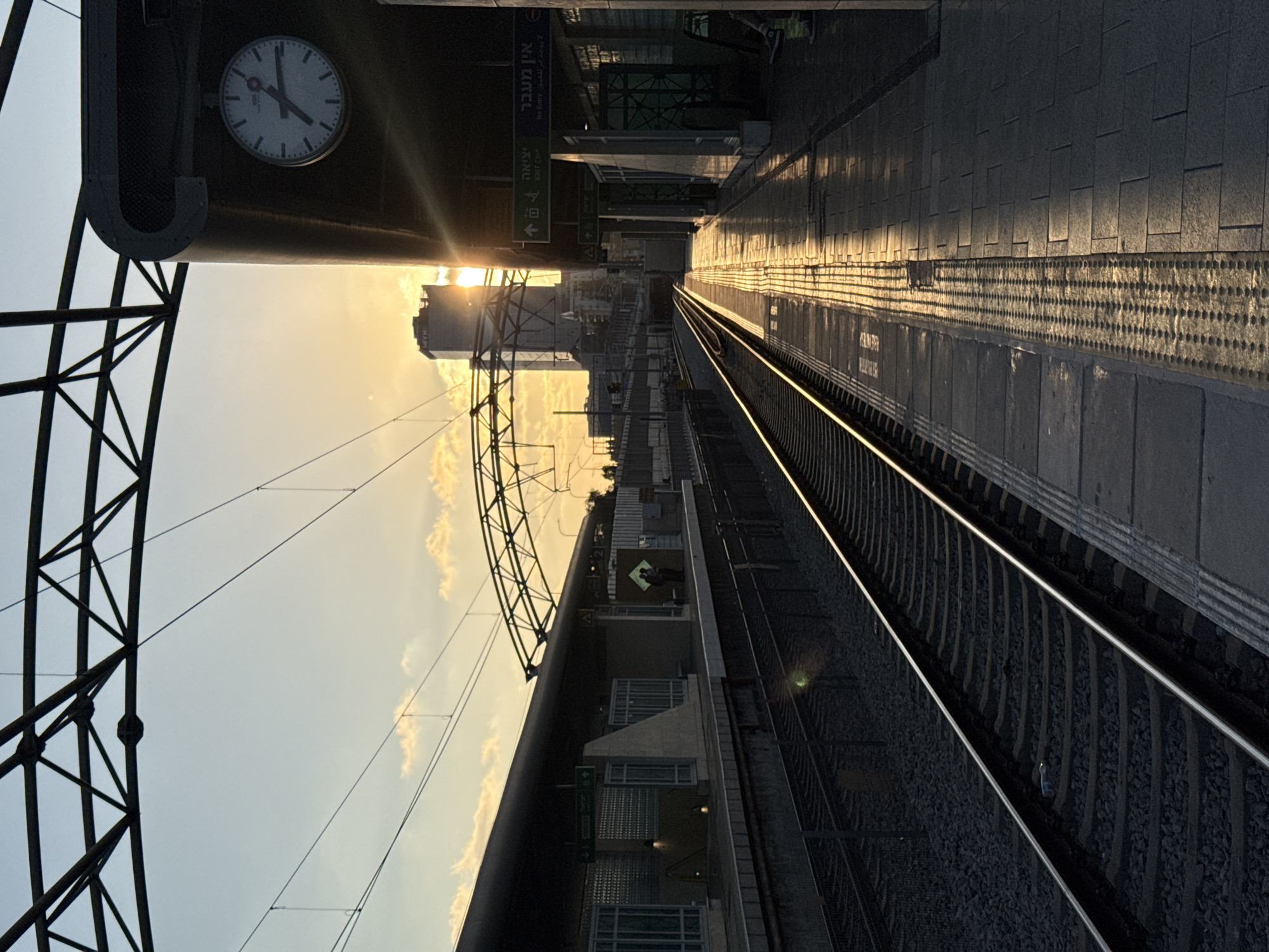 The image depicts a train station platform in Hod HaSharon, Israel, during sunset. The sun is low on the horizon, casting long shadows and a warm golden light across the scene. On the left, train tracks run parallel to the platform, which is lined with textured gray tiles. Overhead, a complex network of metal structures supports the train station's roof and electrical lines. A tall building, partially obscured by the setting sun, rises in the distance. A round clock hangs on the side of the station building on the right, showing the time as approximately 7:15. Signs in Hebrew and English indicate "EXIT ONLY" and "No Entry." A few people are visible on the platform, one sitting and another standing with their back to the camera. The overall atmosphere is serene, with the setting sun creating a dramatic and beautiful contrast with the urban infrastructure.