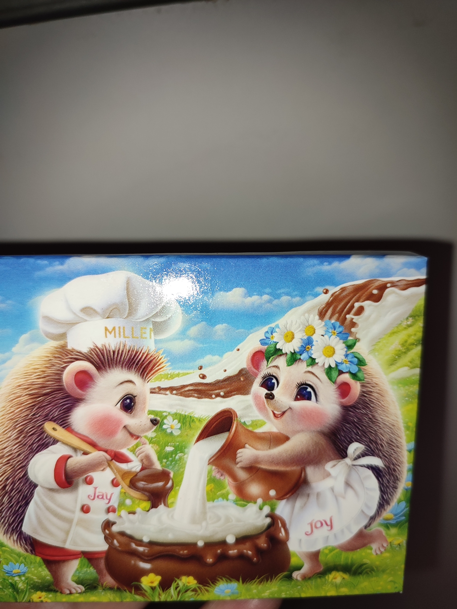The image depicts an illustration of two anthropomorphic hedgehogs in a grassy meadow under a blue sky with white clouds. The hedgehog on the left, wearing a chef's hat with the word "MILLEN" on it and a chef's uniform with red trim, is holding a wooden spoon and appears to be mixing something in a brown bowl. The name "Jay" is embroidered on its uniform. The hedgehog on the right, adorned with a flower crown, is wearing a white apron with the word "Joy" embroidered on it. It is pouring liquid, likely milk, from a jug into the same brown bowl. The bowl is overflowing with a white frothy substance, and chocolate is depicted as flowing down a slope in the background. The image is presented in a somewhat close-up view. There is no information or visual context in the image that would indicate its location within the city of Voskobiinyky, Ukraine.