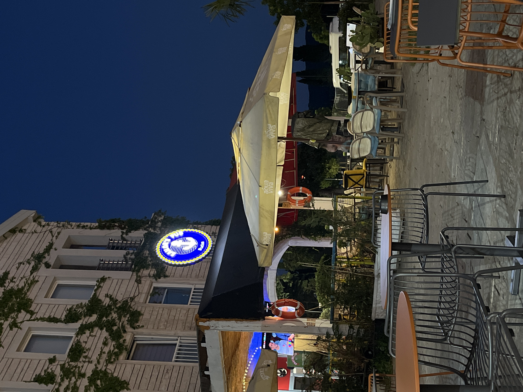 This image depicts an outdoor seating area of a establishment in Budva, Montenegro. The scene is set at dusk, with the sky a deep navy blue. A building with light-colored siding and ivy growing on it serves as the backdrop. A prominent circular neon sign with an anchor motif and the words "Sailor's Pub" is visible on the building.
In the foreground and midground, there are numerous tables and chairs arranged for outdoor dining. Some tables have orange tops, while others are round and appear to be made of wood. The chairs are a mix of designs, including metal wire chairs and upholstered chairs in light blue and cream colors. Several large umbrellas, some a pale yellow and others red, provide shade.
A television screen is visible under an awning, displaying an image of a person. Two life preservers are hanging on the wall near the entrance to the establishment. In the background, there are trees and mountains silhouetted against the sky, and a white van is parked. The ground is paved with grey stones. The overall atmosphere suggests a relaxed evening setting at a pub or restaurant.
