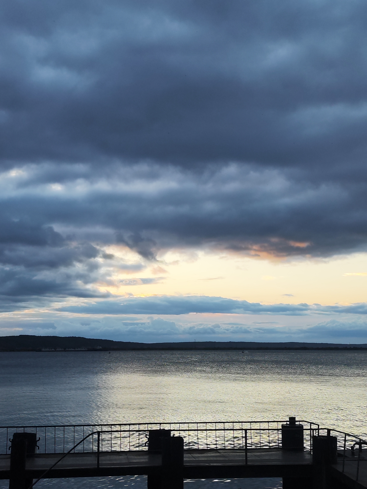 This media file depicts a serene, twilight scene of a body of water, likely a reservoir or large river, viewed from a pier. The sky is filled with dramatic, heavy clouds, with patches of warm, golden light breaking through near the horizon, suggesting either sunrise or sunset. The water reflects the sky's colors and the faint light, creating a shimmering effect. In the foreground, a dark pier with a metal railing and mooring bollards is visible. Beyond the water, a distant, dark landmass forms the horizon, with faint silhouettes of structures visible on it. The overall mood is tranquil and slightly melancholic, perhaps due to the overcast conditions and the subdued light. No people or specific activities are apparent; the focus is on the atmospheric landscape. The location is identified as Chaikovsky, Russia.