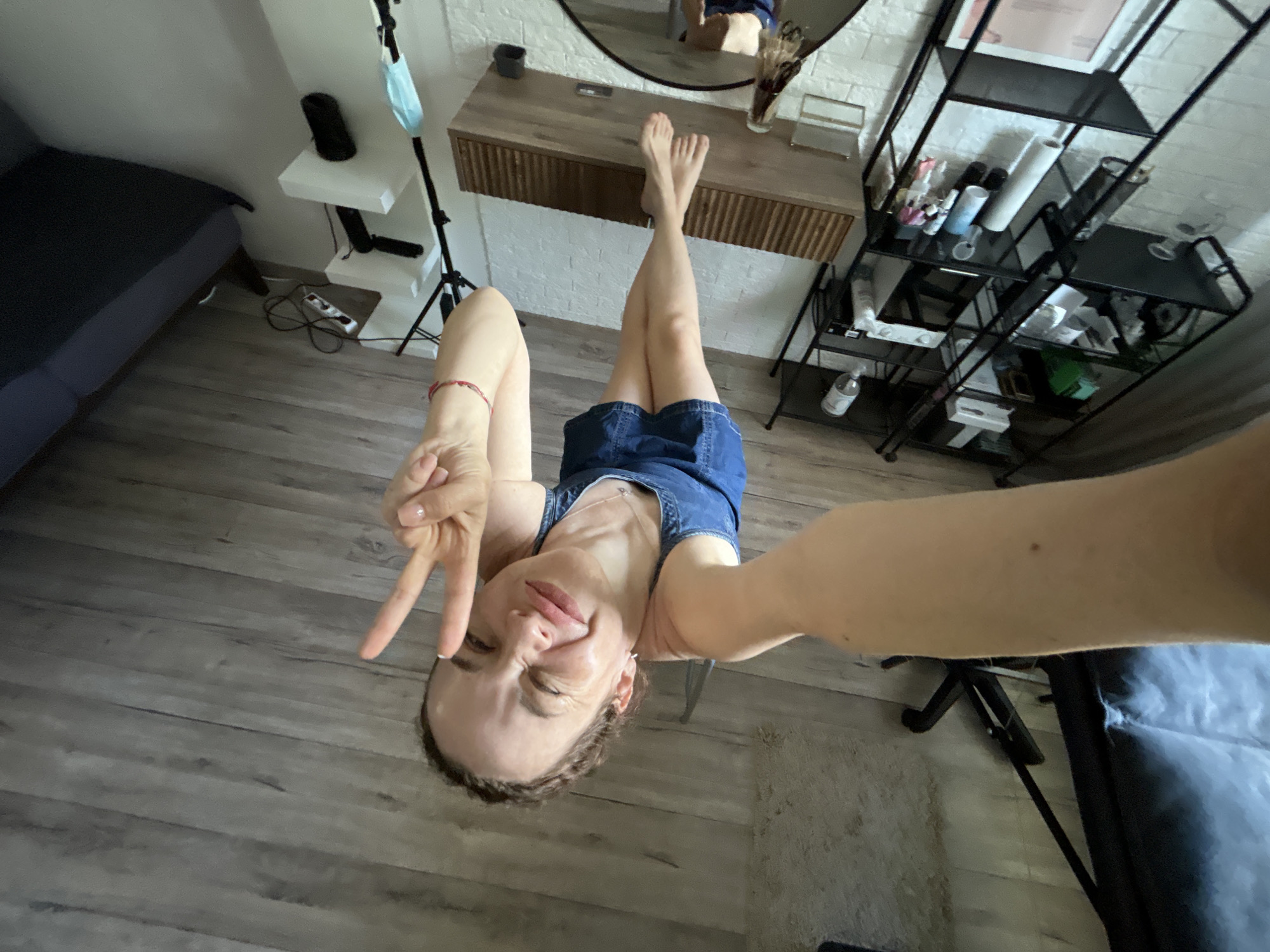 A young woman poses for a selfie in a room. She is positioned upside down with her legs extended upward towards a wall-mounted vanity. Her feet are visible in the reflection of a round mirror above the vanity. She's wearing a sleeveless blue denim romper. Her left arm is extended out with her hand making a peace sign, while her right arm extends out holding the camera. To the left of the vanity is a white shelf with a black cylinder on top and a face mask hanging from a tripod stand. To the right of the vanity is a black metal shelving unit filled with various containers. The room has light grey wood-look flooring. A bed with a dark blue comforter is visible in the background. A chair with a black metal frame and a dark blue seat is visible in the right corner. There are no indications in the image of the location being Chaikovsky, Russia.