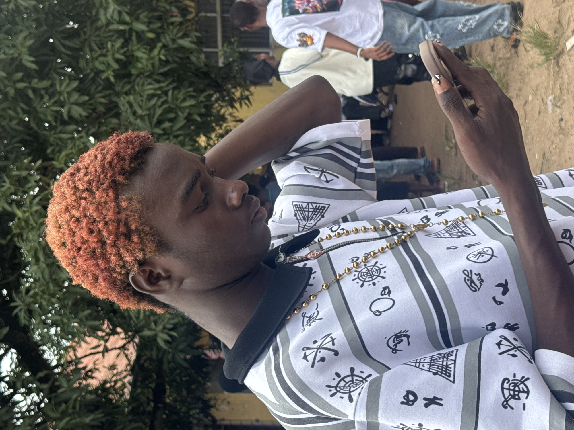 The image shows a young Black male with short, reddish-orange curly hair looking down at a smartphone he is holding in his right hand. He is wearing a white collared shirt with a black collar, adorned with a pattern of black symbols and horizontal grey and black stripes. A gold chain with round beads is visible around his neck. His right hand, which is holding the phone, has manicured fingernails with black and white designs. He is standing outdoors, with his left hand resting on the back of his head.
In the background, other people are visible, appearing to be gathered in an outdoor area with some buildings and trees. The ground appears to be dirt or sand. The overall atmosphere suggests a casual gathering or event. The location is identified as Ihiagwa, Nigeria.