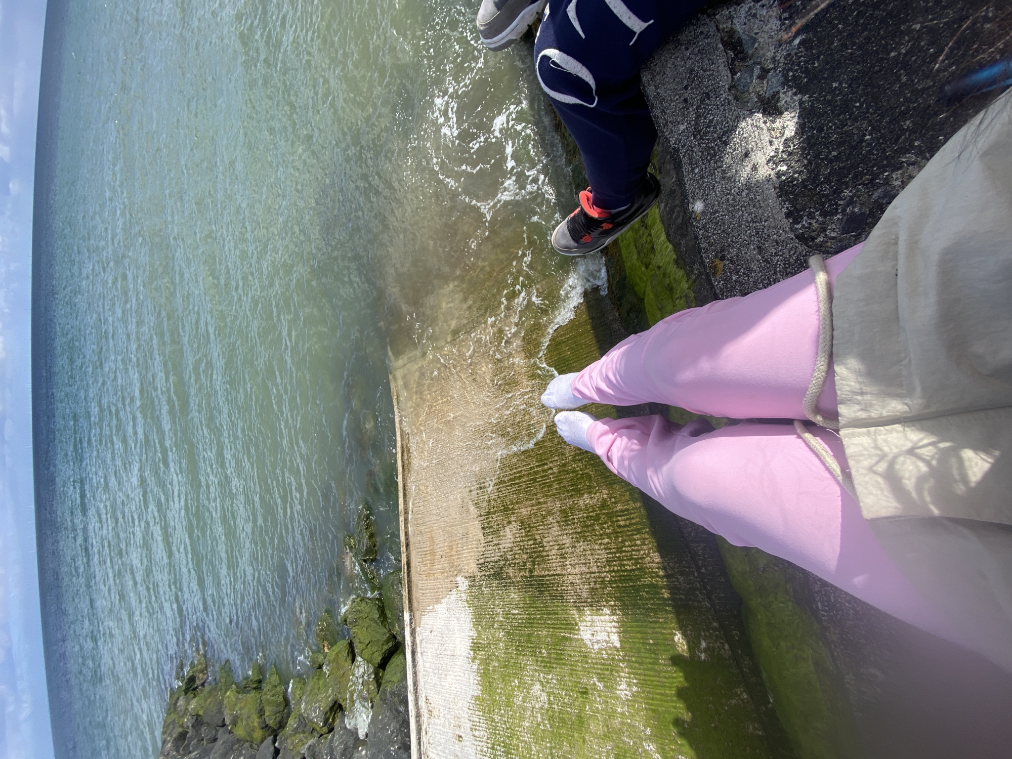 The image shows two people sitting on a mossy, concrete barrier at the edge of a body of water, likely the sea. One person is wearing navy blue sweatpants with white lettering and grey shoes. The other person is wearing pink leggings with a white jacket. Water is lapping against the barrier. The scene is Old Colwyn, United Kingdom, specifically a coastal area.